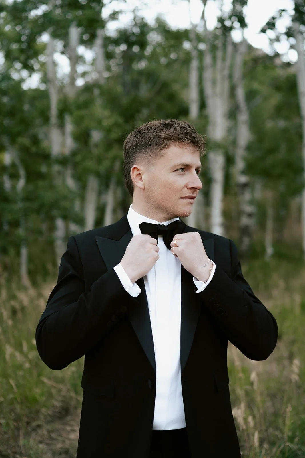 A groom in a black tuxedo from Saint Laurent prepares himself to see his bride for the first time amid a sunlit grove of aspen trees - Poise and Ivy Imagery