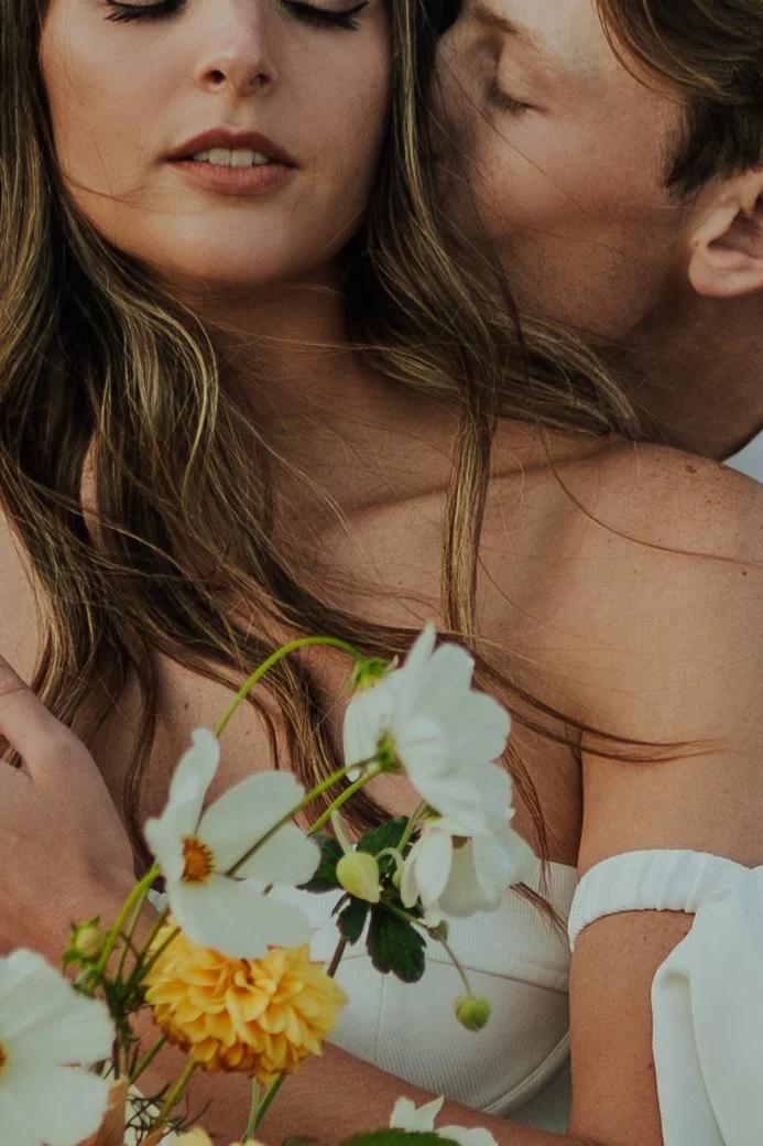 A bride and groom in a wedding gown and a tan suit stand in a meadow with mountains and a lake behind them - photography by Poise and Ivy Imagery