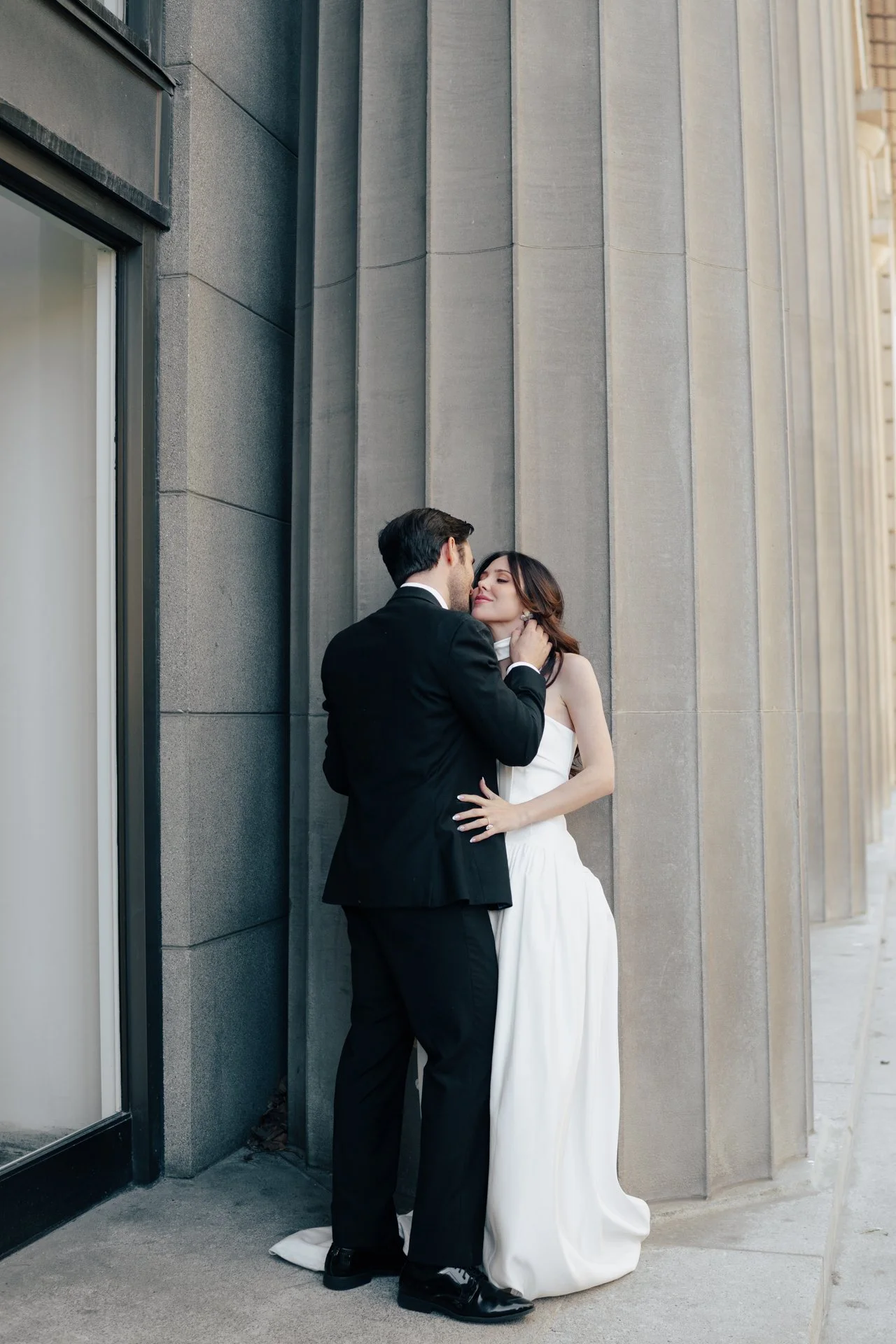 A bride and groom stand together among roman - style architecture in an elegant wedding gown and a classic black suit