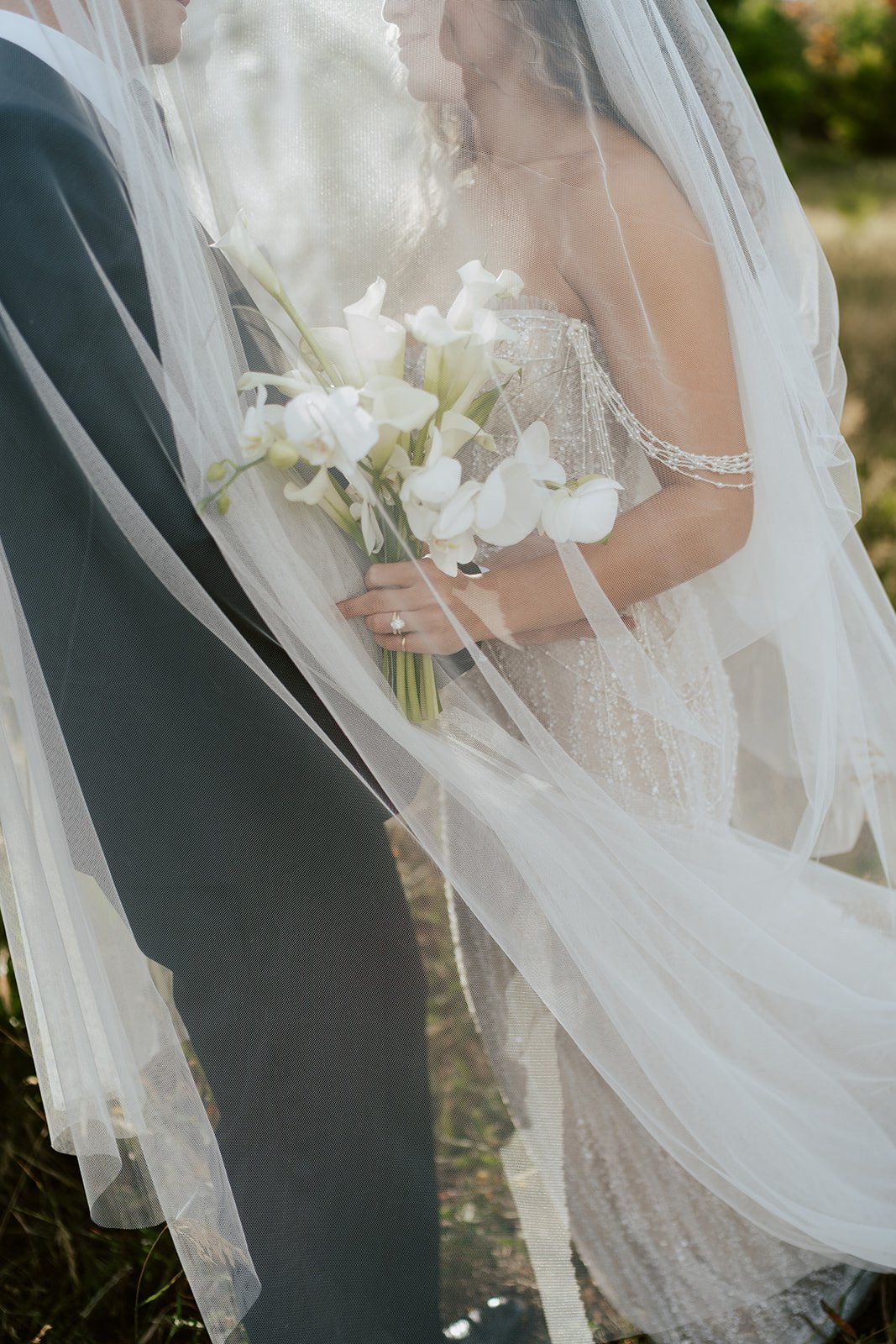 a bride and a groom stand together under a long bridal veil in a lush aspen grove. the bride wears a beaded wedding gown and the groom wears a tuxedo. Photography by Poise and Ivy Imagery