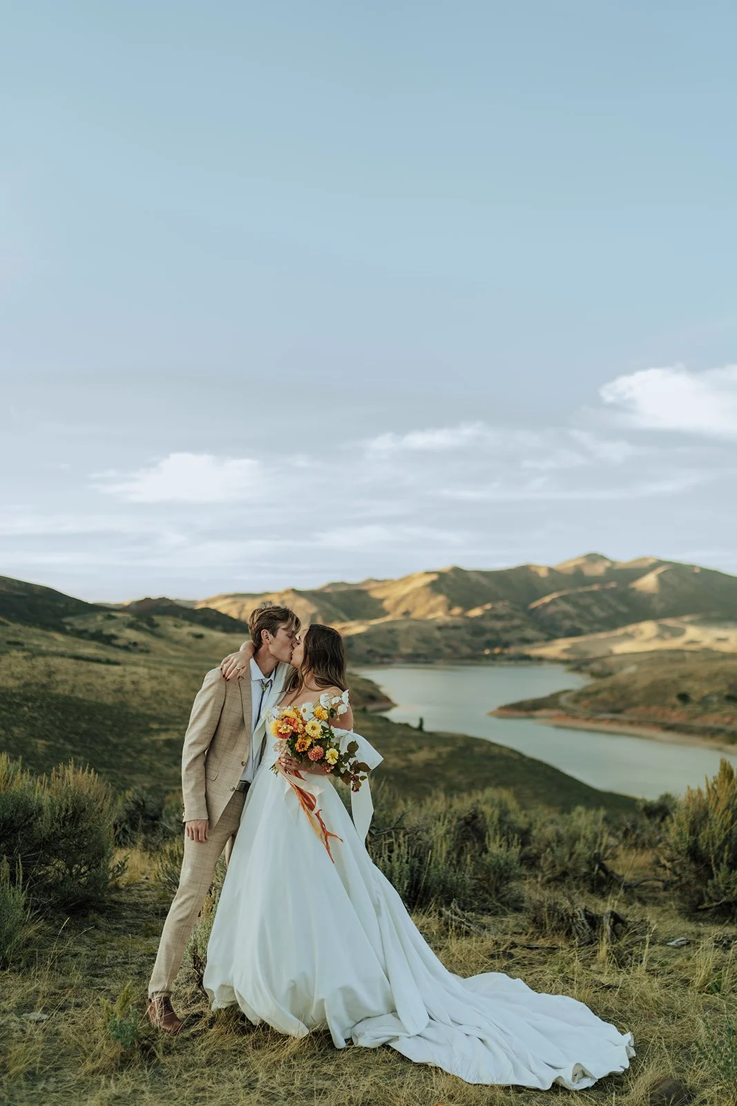 A bride and groom in a wedding gown and a tan suit stand in a meadow with mountains and a lake behind them - photography by Poise and Ivy Imagery