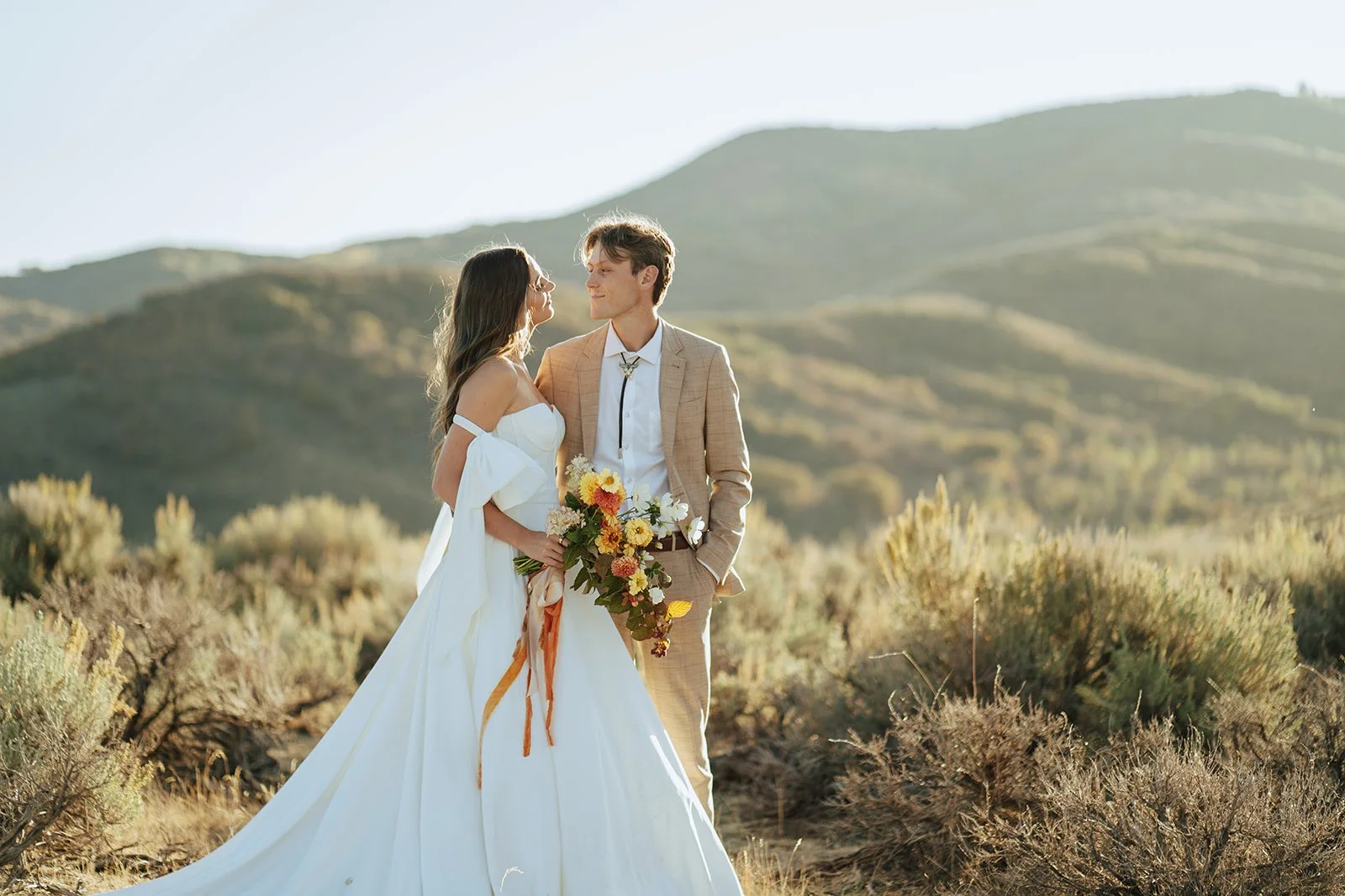 A bride and groom in a wedding gown and a tan suit stand in a meadow with mountainous peaks behind them - photography by Poise and Ivy Imagery
