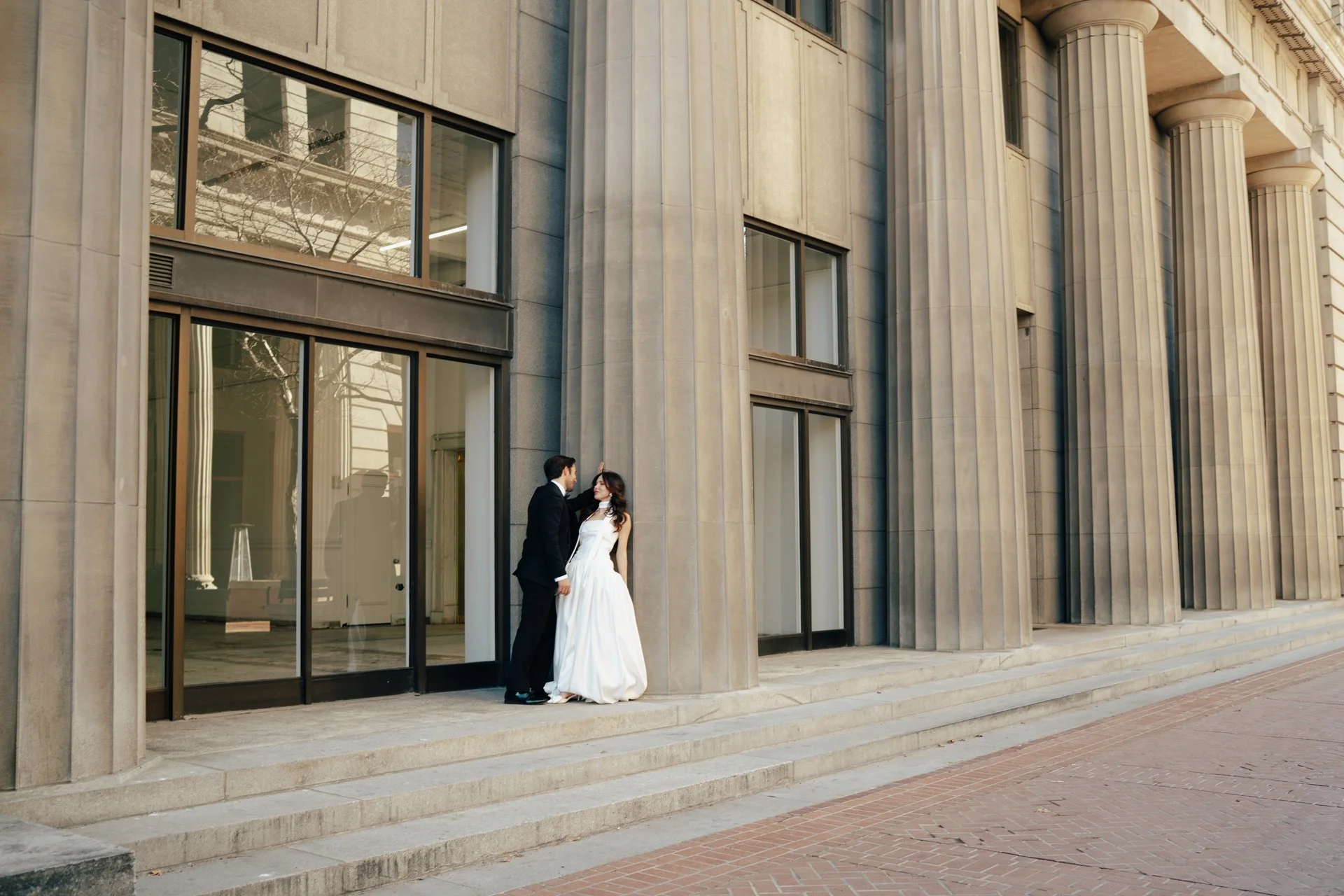 A bride and groom stand together among roman - style architecture in an elegant wedding gown and a classic black suit