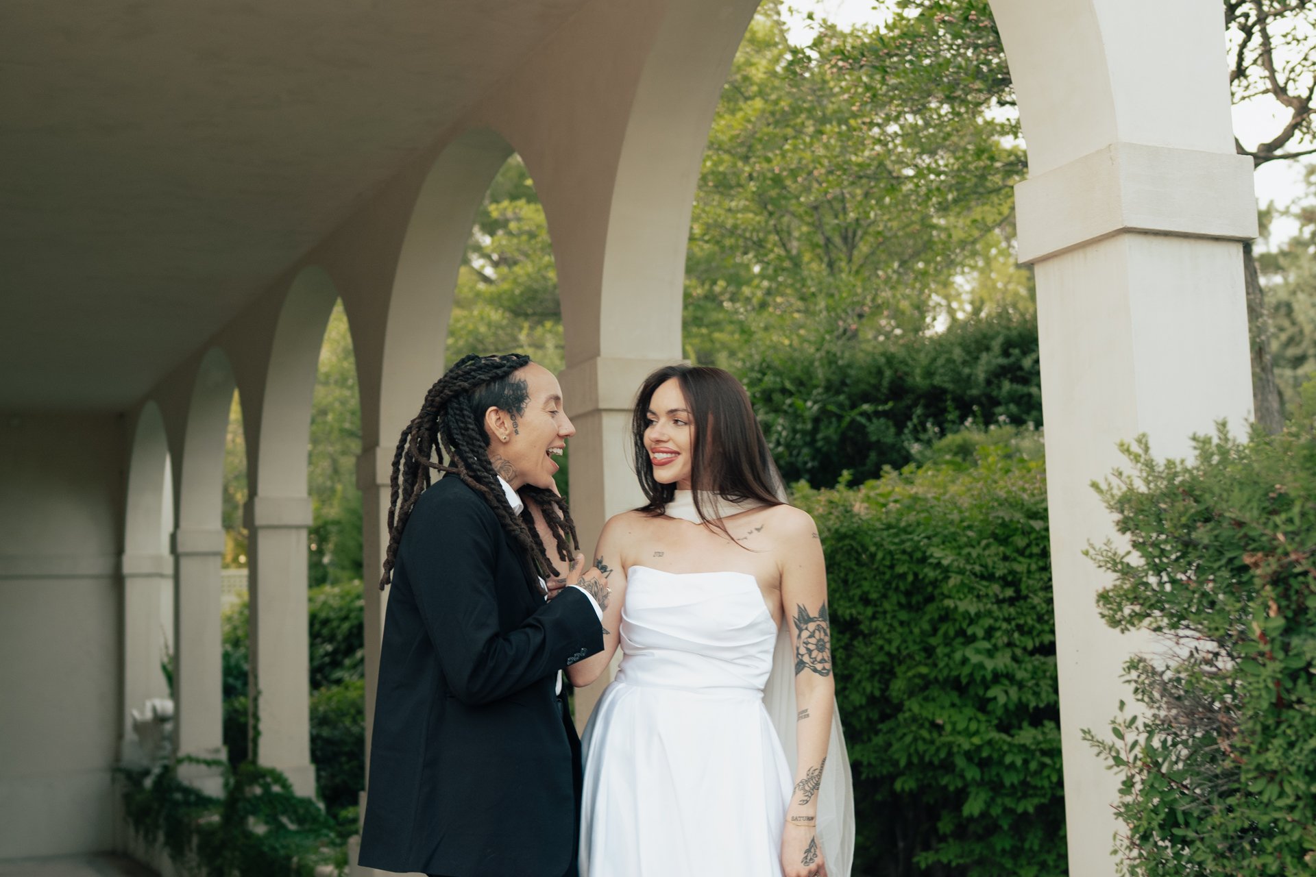 Two brides in wedding attire embrace under an ornate outdoor arched corridor - photographed by Poise and Ivy Imagery