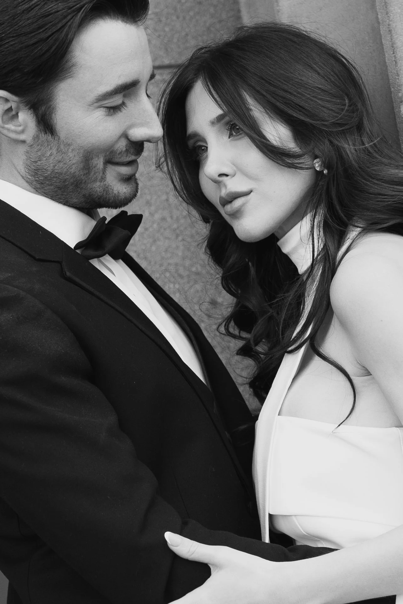 A bride and groom stand together among roman - style architecture in an elegant wedding gown and a classic black suit