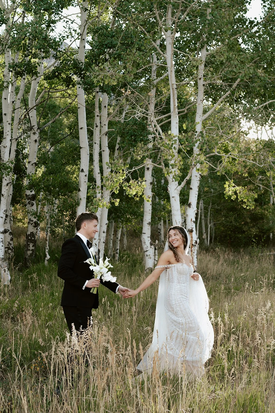 a bride and a groom walk together in a lush aspen grove. the bride wears a beaded wedding gown with a bridal veil and the groom wears a tuxedo. Photography by Poise and Ivy Imagery