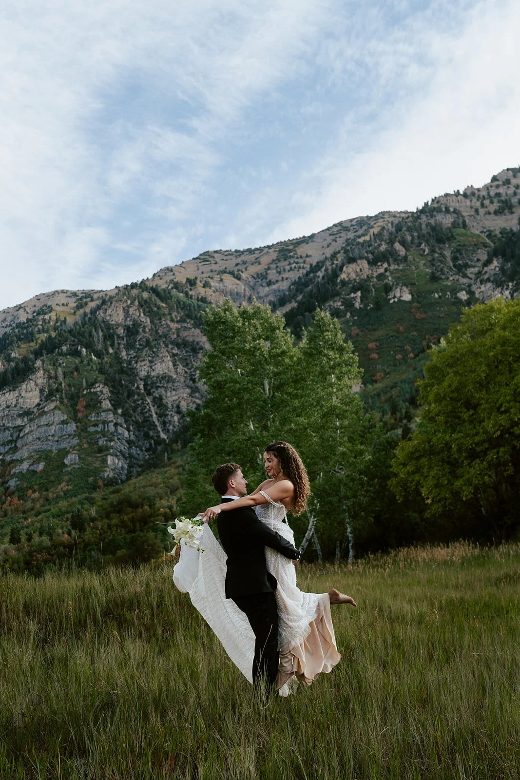 a bride and groom stand together in a softly sunlit meadow, wearing a beaded wedding gown and a black tuxedo. Photography by Poise and Ivy Imagery