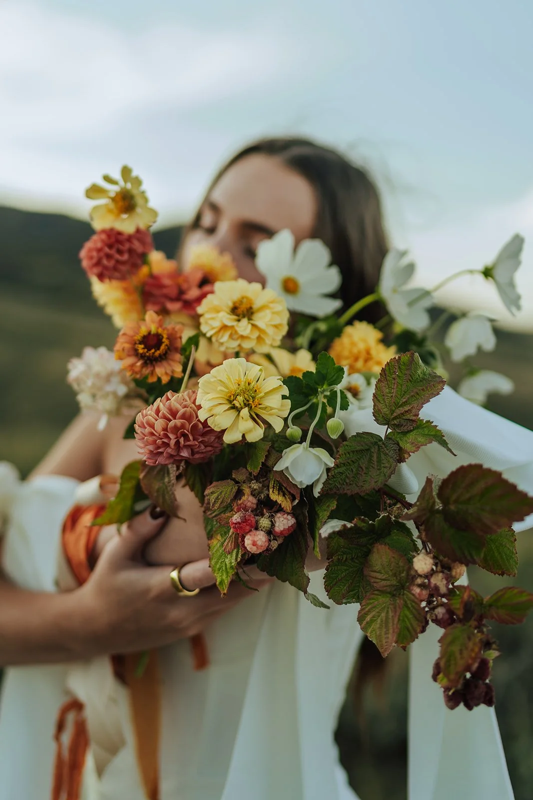 A bride holds a bouquet of red, orange, yellow, and white blooms up to her face. Photography by Poise and Ivy Imagery