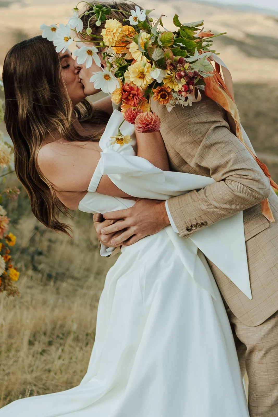 A bride and groom in a wedding gown and a tan suit stand in a meadow with mountains and a lake behind them - photography by Poise and Ivy Imagery
