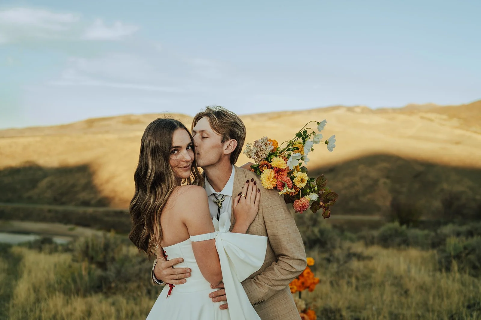 A bride and groom in a wedding gown and a tan suit stand in a meadow with mountains and a lake behind them - photography by Poise and Ivy Imagery