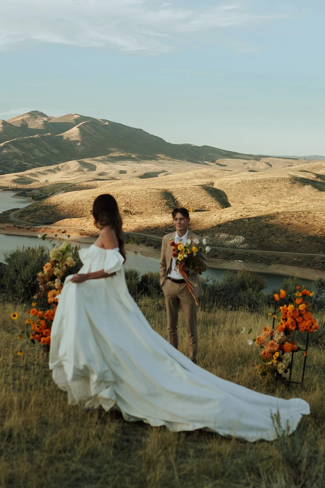 A bride and groom in a wedding gown and a tan suit stand in a meadow with mountains and a lake behind them - photography by Poise and Ivy Imagery