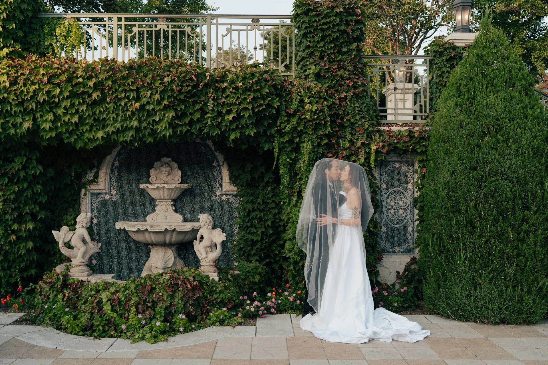 Two brides in wedding attire embrace under a wedding veil next to an ivy covered wall and an ornate fountain - photographed by Poise and Ivy Imagery