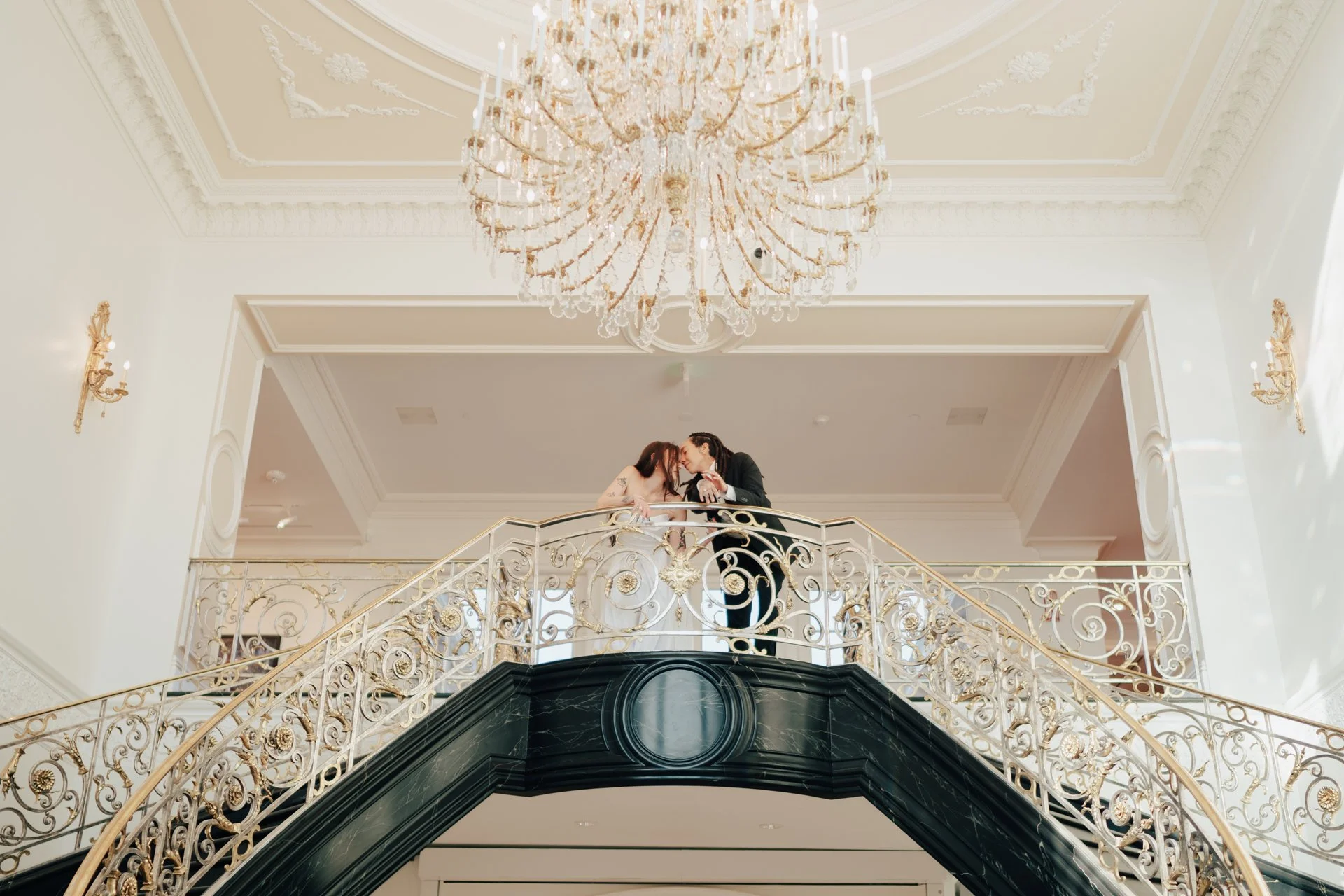 two brides embrace on an ornate double staircase - poise and ivy imagery