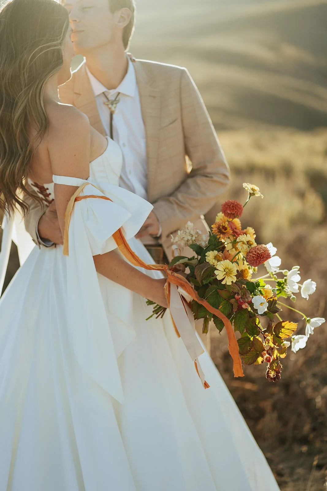A bride and groom in a wedding gown and a tan suit stand in a meadow with mountainous peaks behind them - photography by Poise and Ivy Imagery