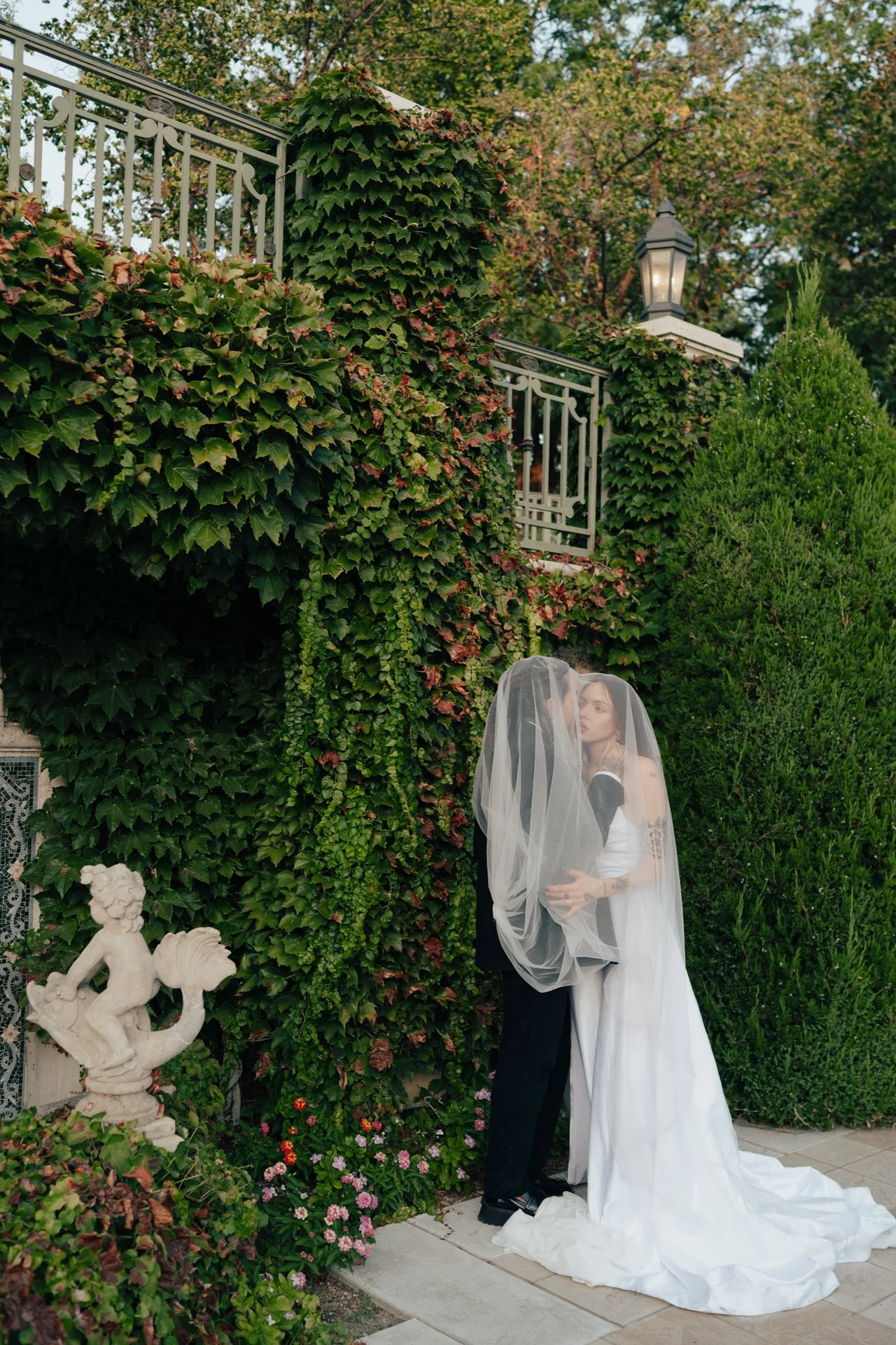 Two brides in wedding attire embrace under an ornate outdoor arched corridor - photographed by Poise and Ivy Imagery