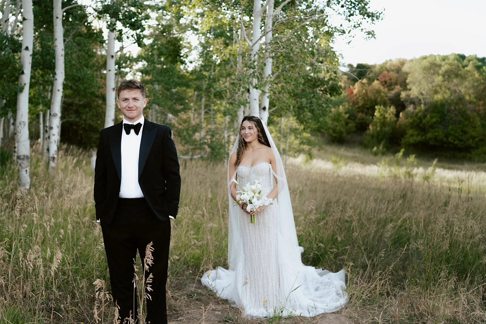 a bride and a groom stand together in a lush aspen grove. the bride wears a beaded wedding gown with a bridal veil and the groom wears a tuxedo. Photography by Poise and Ivy Imagery