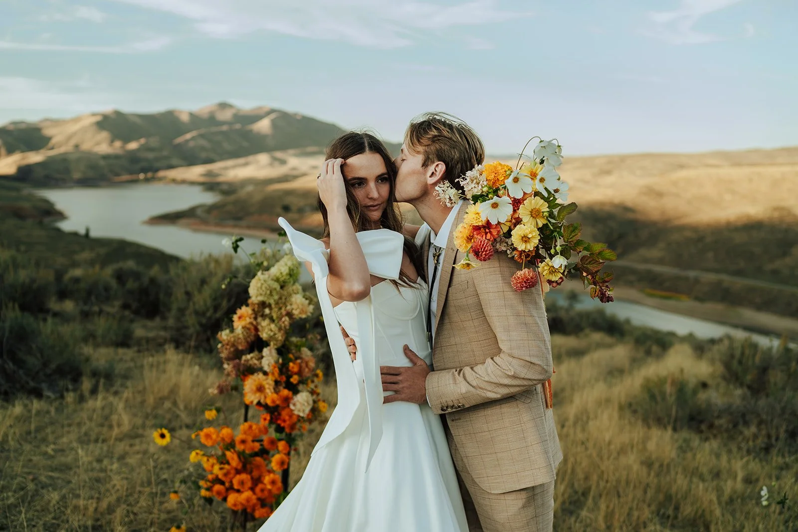 A bride and groom in a wedding gown and a tan suit stand in a meadow with mountains and a lake behind them - photography by Poise and Ivy Imagery