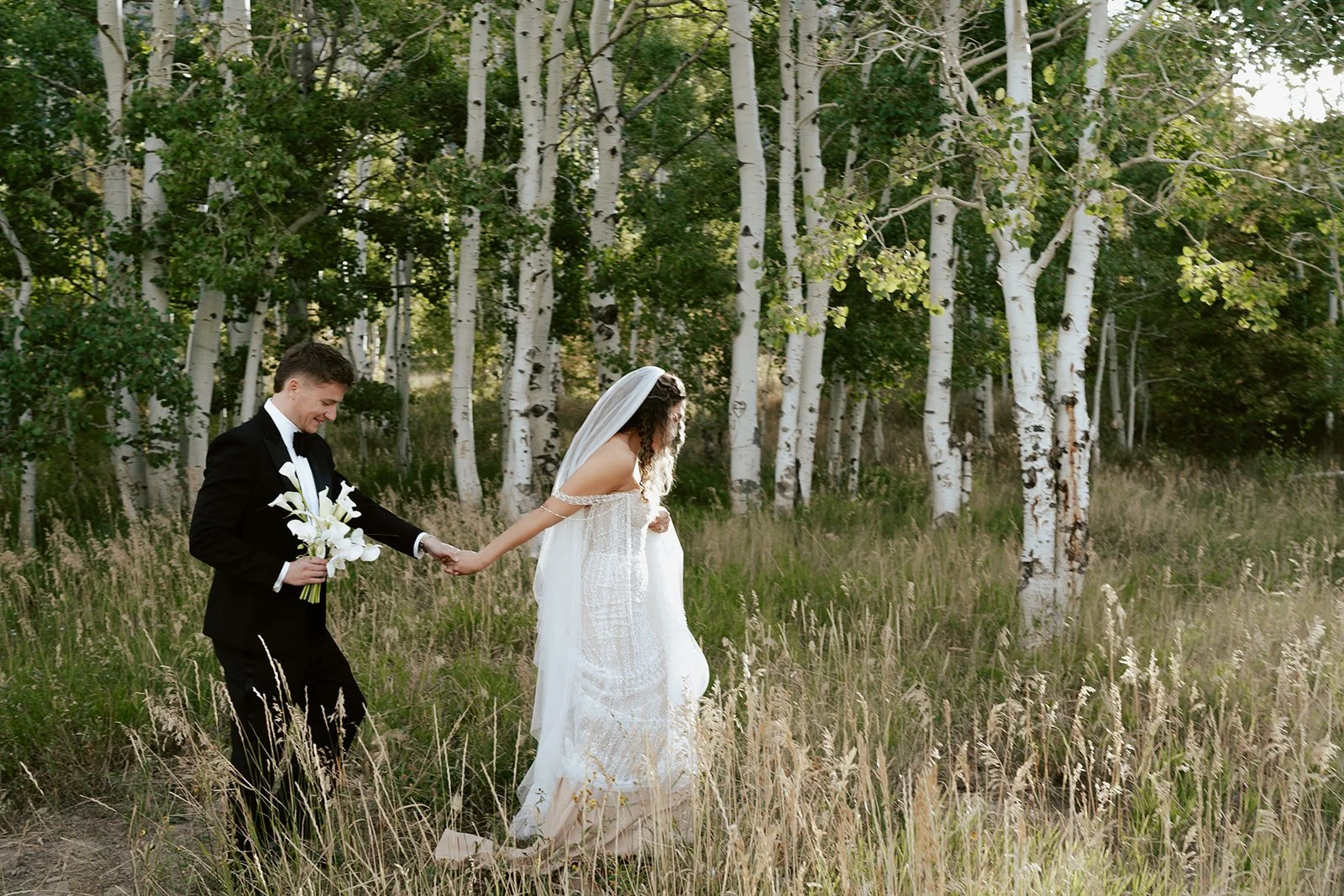 a bride and a groom walk together in a lush aspen grove. the bride wears a beaded wedding gown with a bridal veil and the groom wears a tuxedo. Photography by Poise and Ivy Imagery