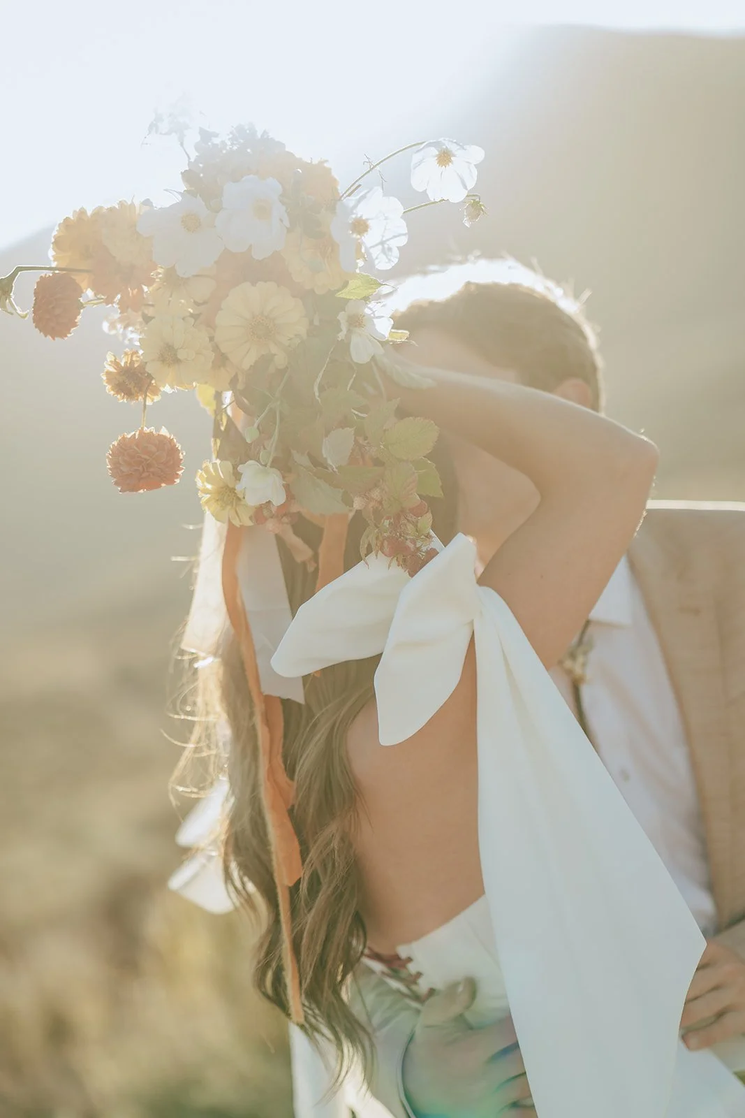 A bride and groom in a wedding gown and a tan suit stand in a meadow with mountainous peaks behind them - photography by Poise and Ivy Imagery