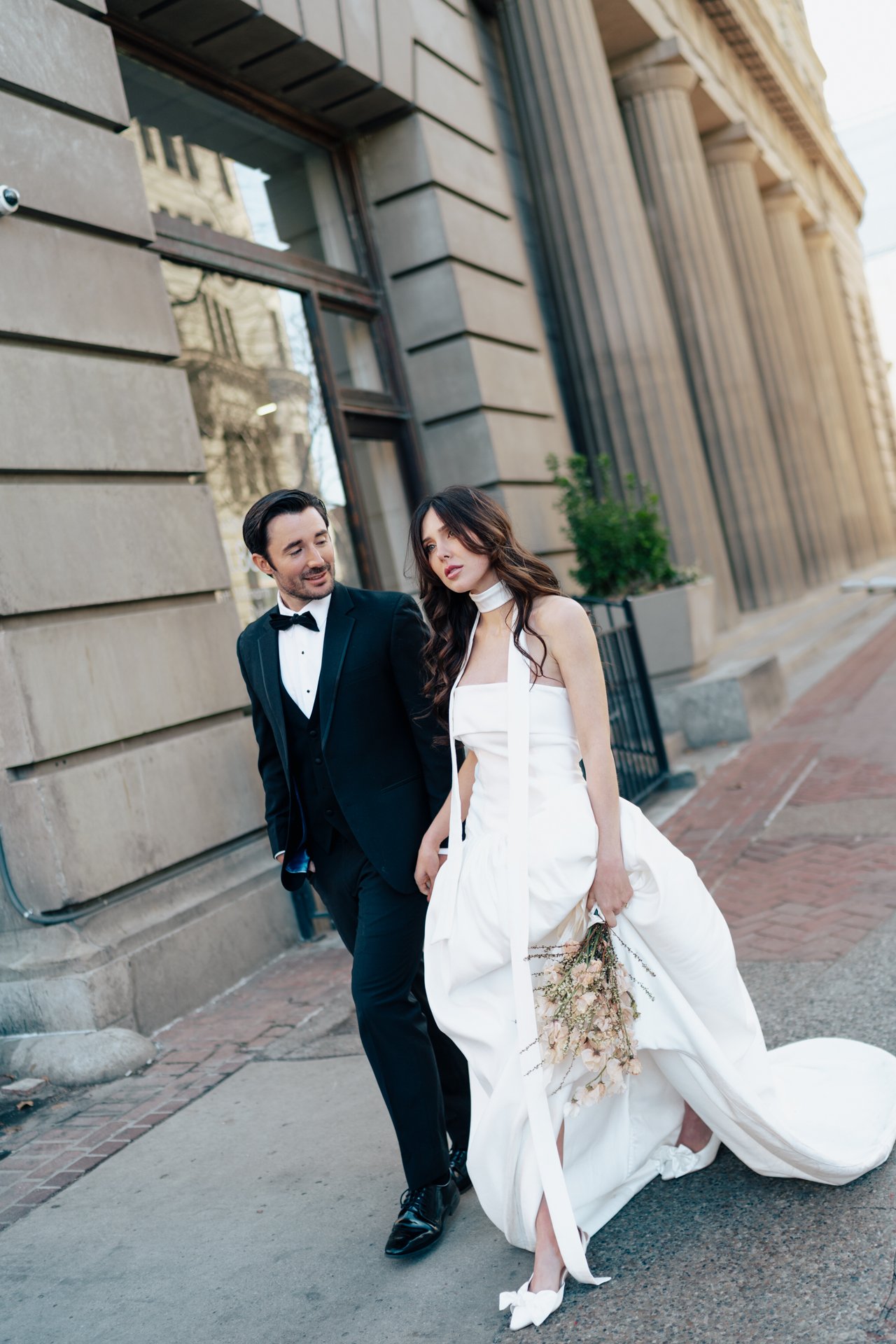 A bride and groom stand together among roman - style architecture in an elegant wedding gown and a classic black suit