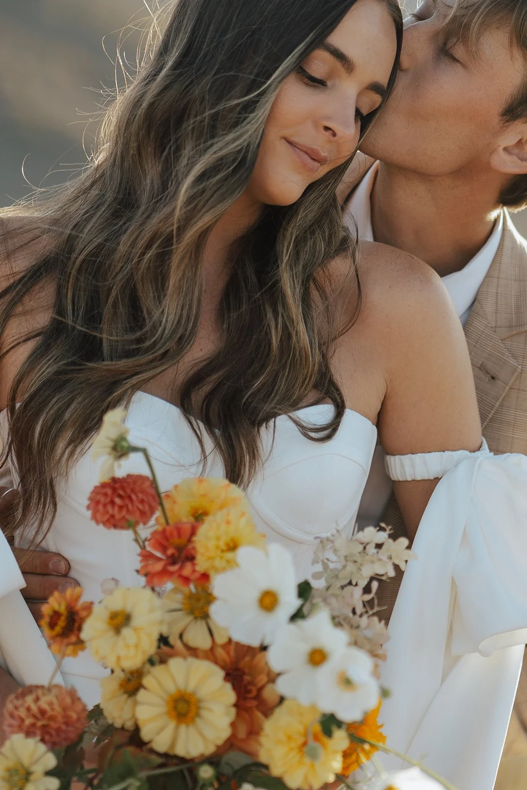A bride and groom in a wedding gown and a tan suit stand in a meadow with mountainous peaks behind them - photography by Poise and Ivy Imagery