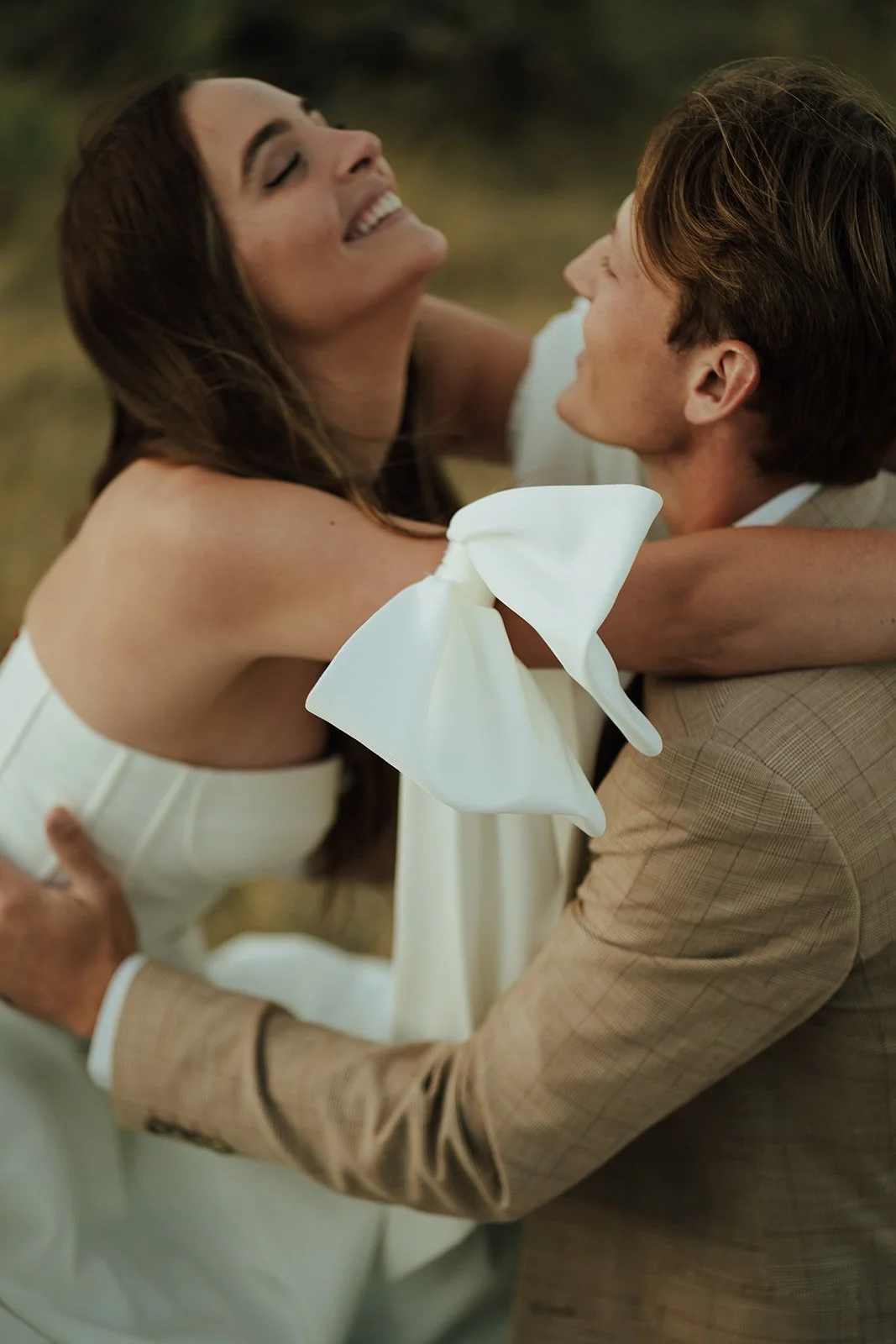 A bride and groom in a wedding gown and a tan suit stand in a meadow with mountains and a lake behind them - photography by Poise and Ivy Imagery