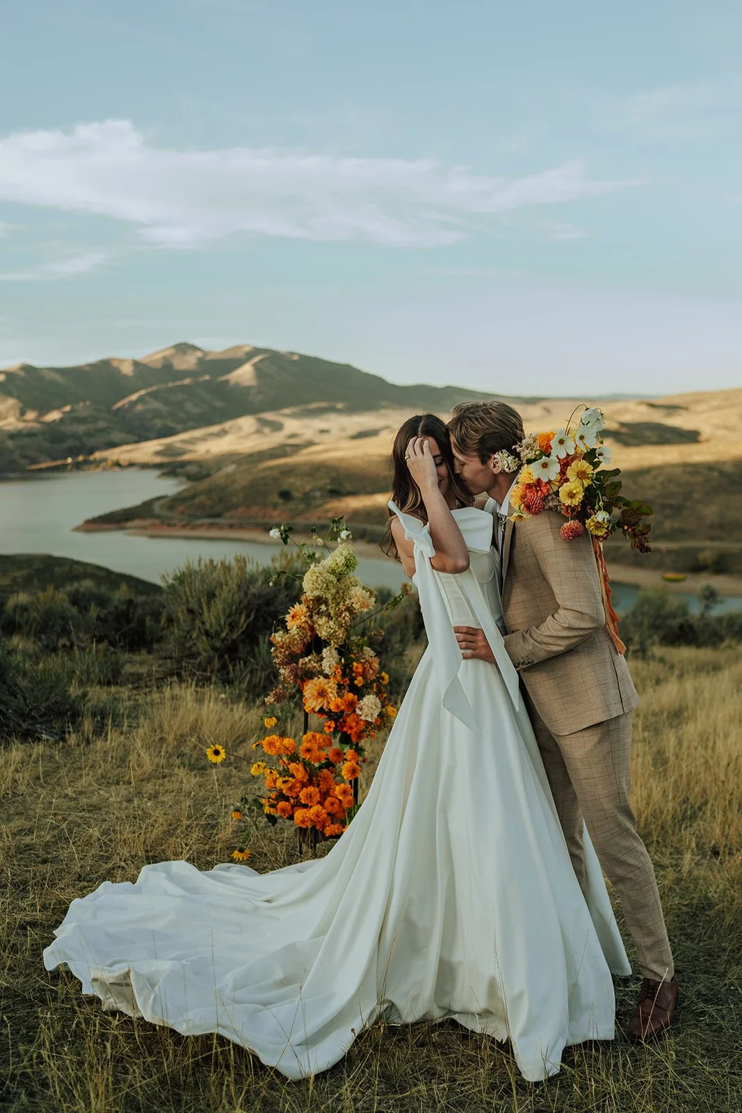 A bride and groom in a wedding gown and a tan suit stand in a meadow with mountains and a lake behind them - photography by Poise and Ivy Imagery