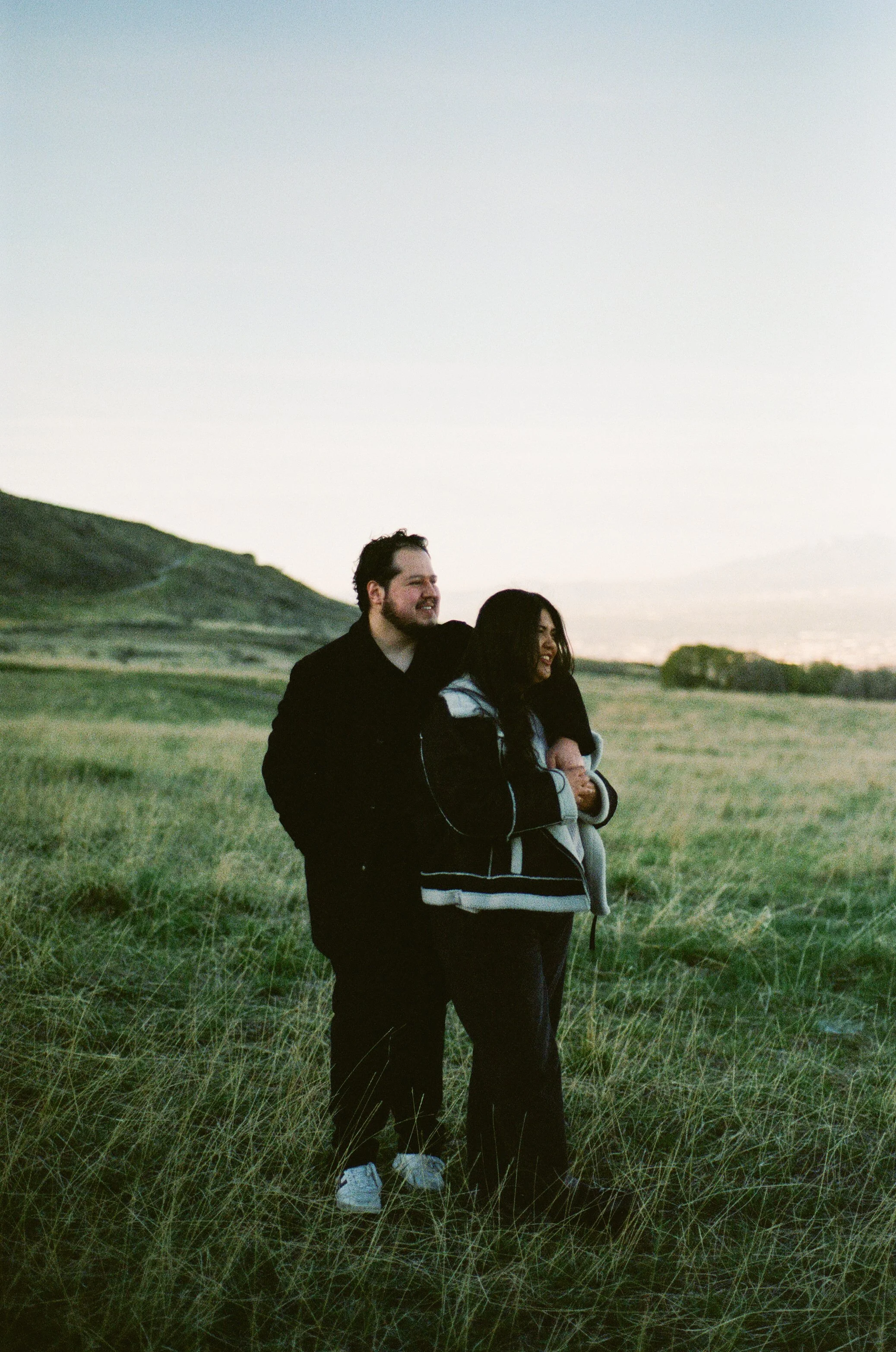 A couple stands together in a softly lit grassy field, photographed on color negative film by Poise and Ivy Imagery