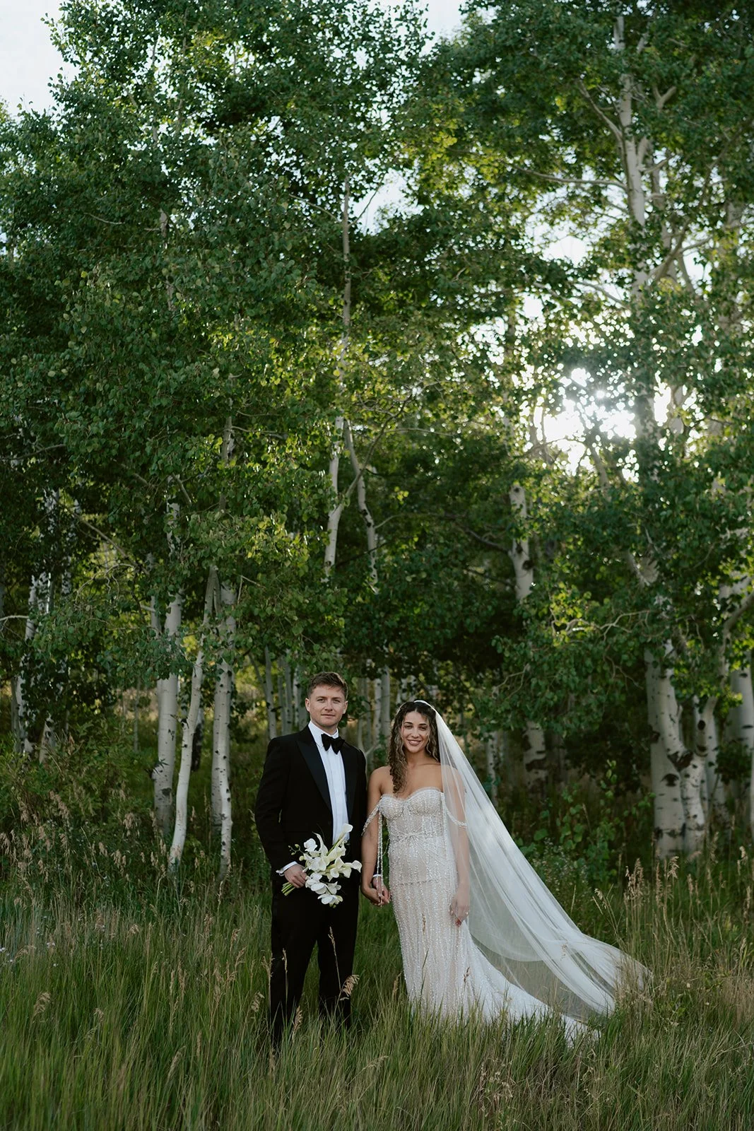 Bride and groom stand in an aspen meadow wearing a pearl beaded wedding gown and a black tuxedo - photo taken by poise and ivy imagery