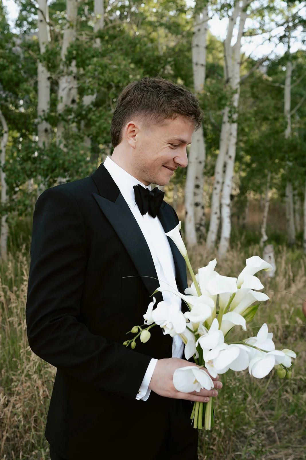 a groom wearing a black tuxedo holds his bride's wedding bouquet made of white calla lilies - photography by poise and ivy imagery