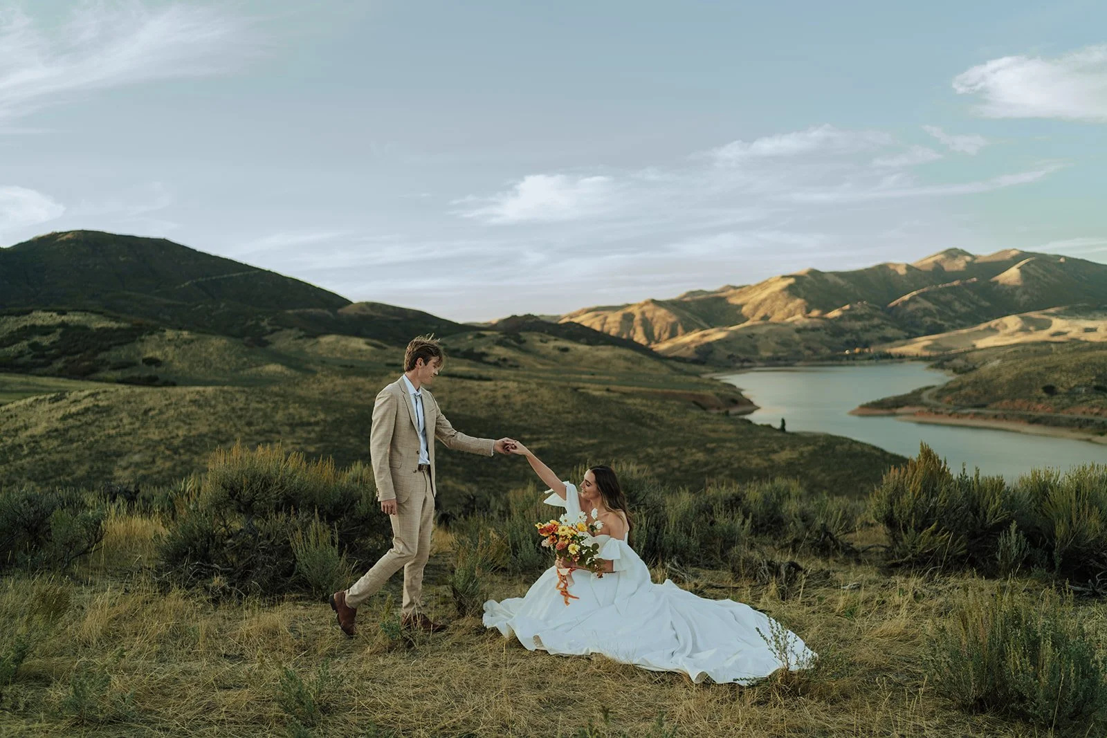A bride and groom in a wedding gown and a tan suit stand in a meadow with mountains and a lake behind them - photography by Poise and Ivy Imagery