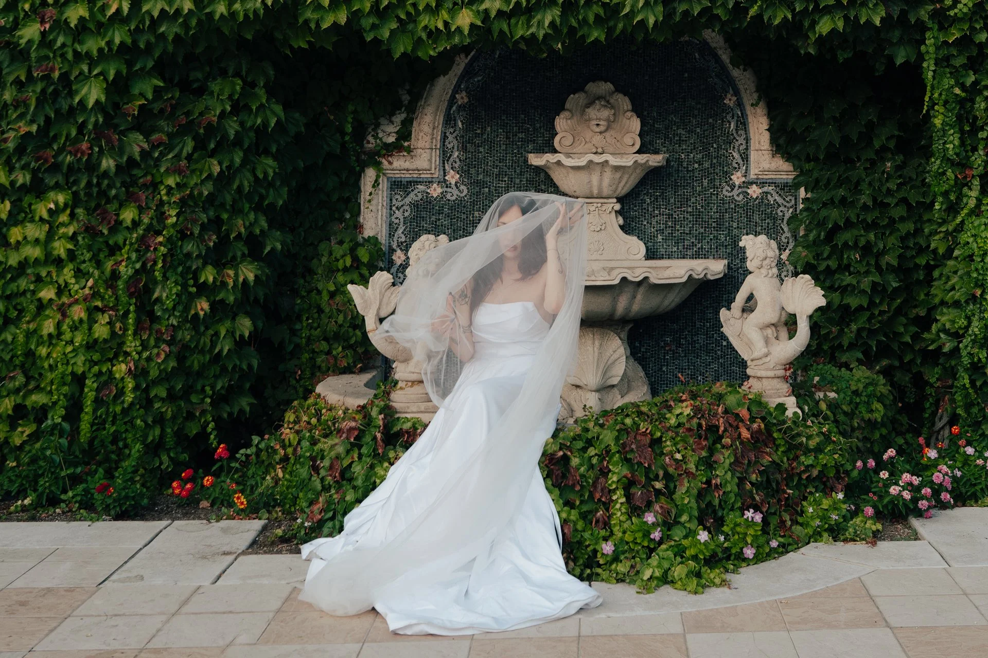 Two brides in wedding attire embrace under a wedding veil next to an ivy covered wall and an ornate fountain - photographed by Poise and Ivy Imagery