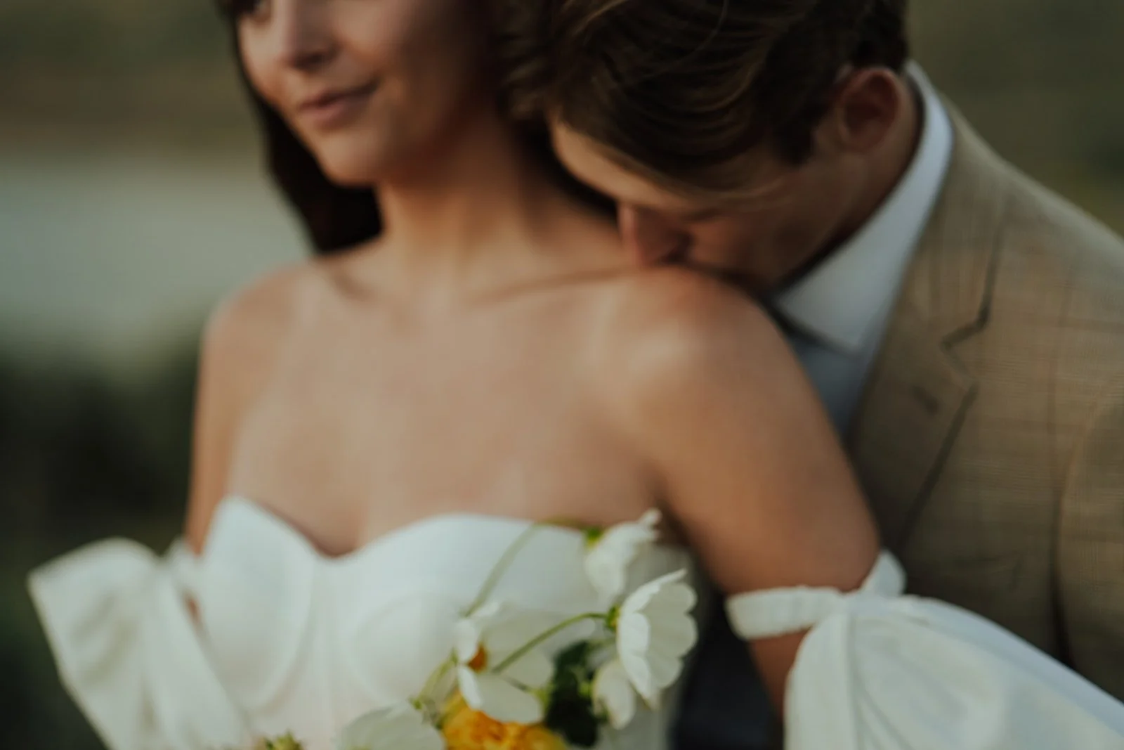 A bride and groom in a wedding gown and a tan suit stand in a meadow with mountains and a lake behind them - photography by Poise and Ivy Imagery