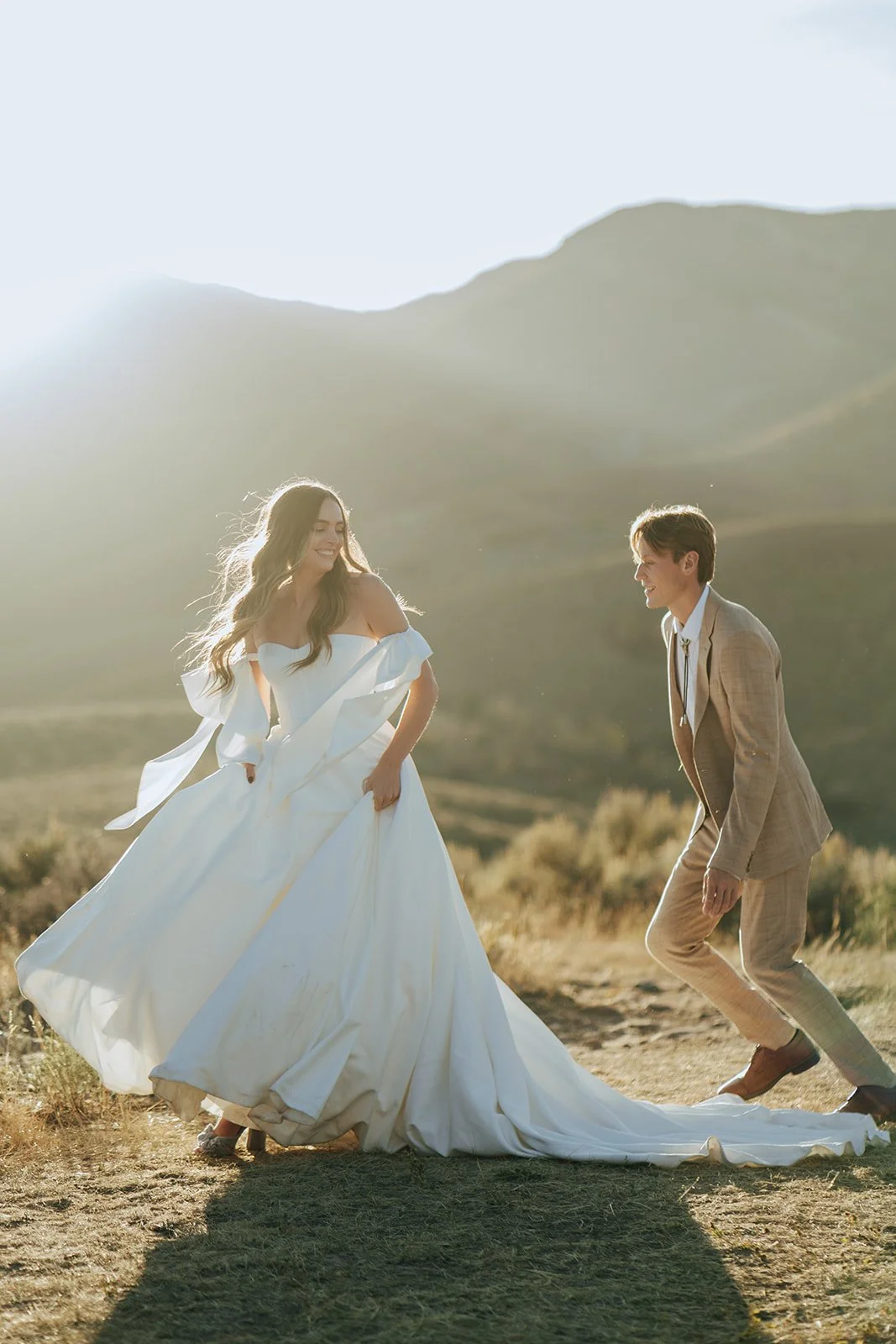 A bride and groom in a wedding gown and a tan suit stand in a meadow with mountainous peaks behind them - photography by Poise and Ivy Imagery