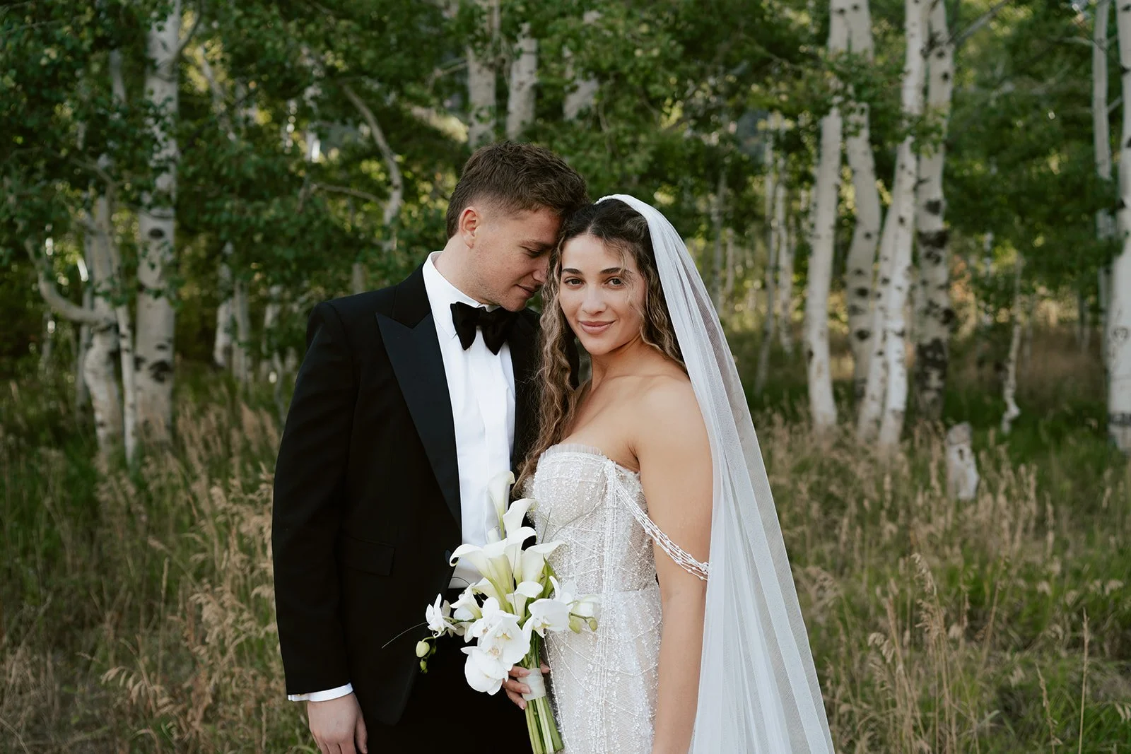 a bride and a groom stand together in a lush aspen grove. the bride wears a beaded wedding gown with a bridal veil and the groom wears a tuxedo. Photography by Poise and Ivy Imagery