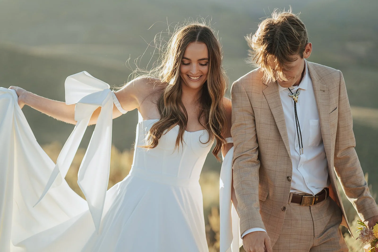 A bride and groom in a wedding gown and a tan suit stand in a meadow with mountainous peaks behind them - photography by Poise and Ivy Imagery