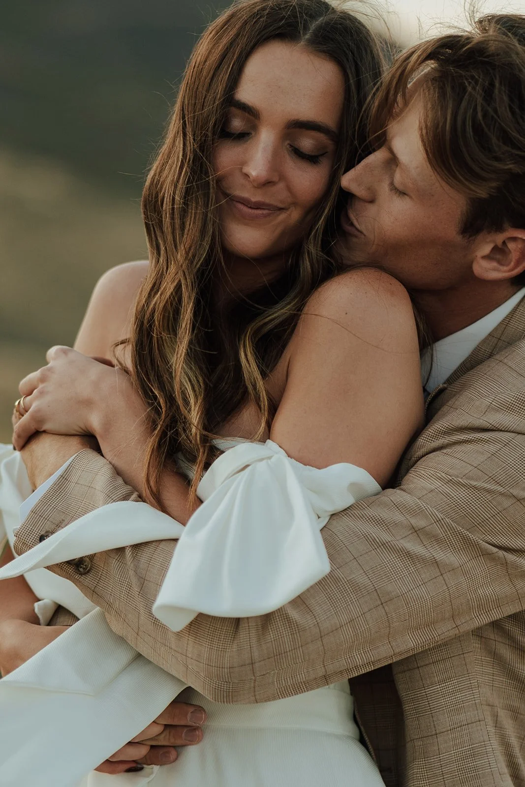 A bride and groom in a wedding gown and a tan suit stand in a meadow with mountains and a lake behind them - photography by Poise and Ivy Imagery