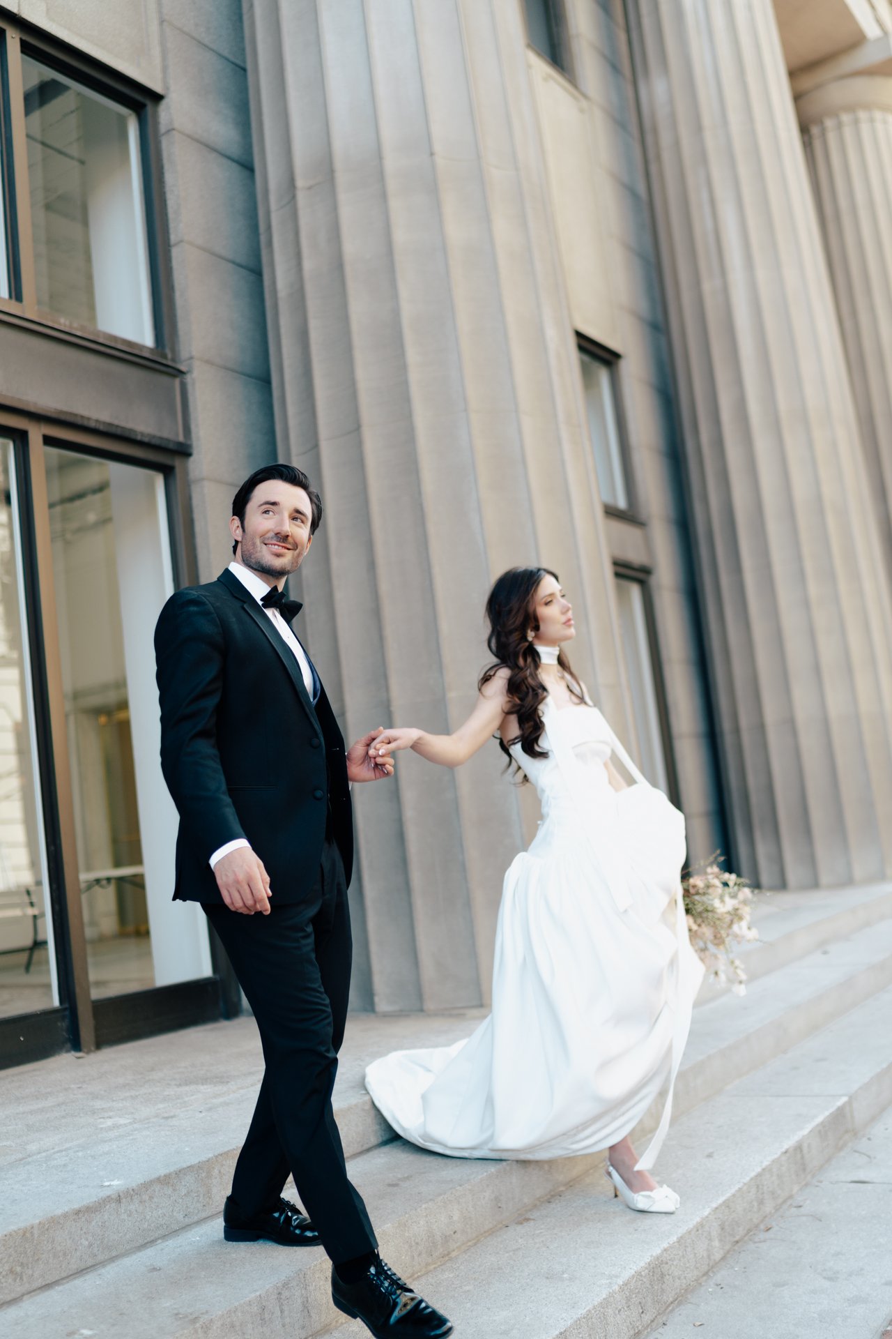 A bride and groom stand together among roman - style architecture in an elegant wedding gown and a classic black suit