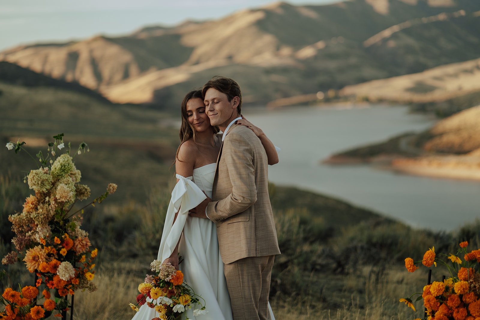 A bride and groom in a wedding gown and a tan suit stand in a meadow with mountains and a lake behind them - photography by Poise and Ivy Imagery