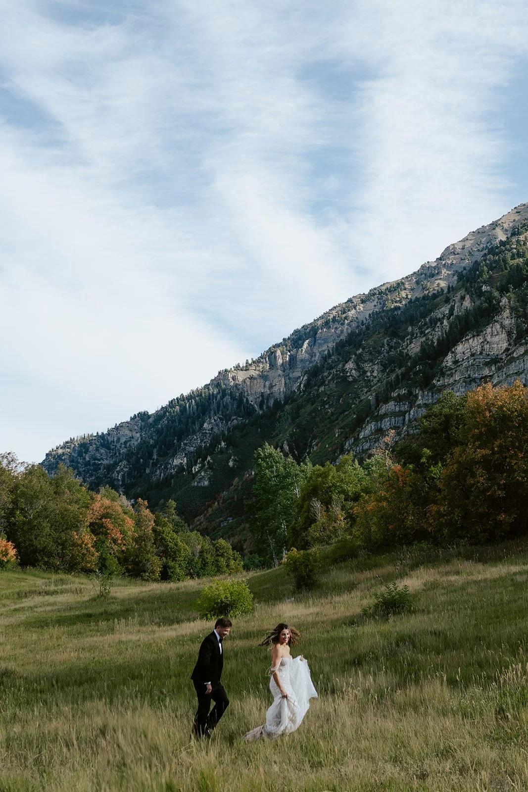 a bride and a groom run together through a sunlit meadow. the bride wears a beaded wedding gown and the groom wears a tuxedo. Photography by Poise and Ivy Imagery