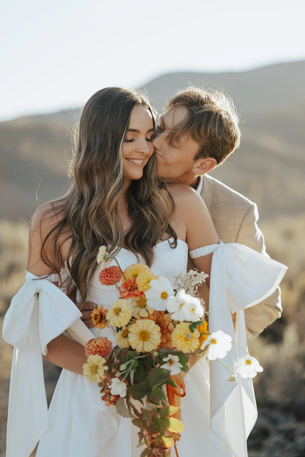 A bride and groom in a wedding gown and a tan suit stand in a meadow with mountainous peaks behind them - photography by Poise and Ivy Imagery