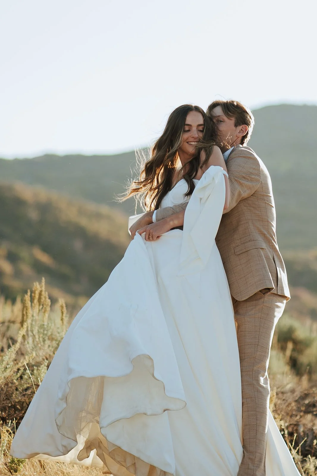 A bride and groom in a wedding gown and a tan suit stand in a meadow with mountainous peaks behind them - photography by Poise and Ivy Imagery