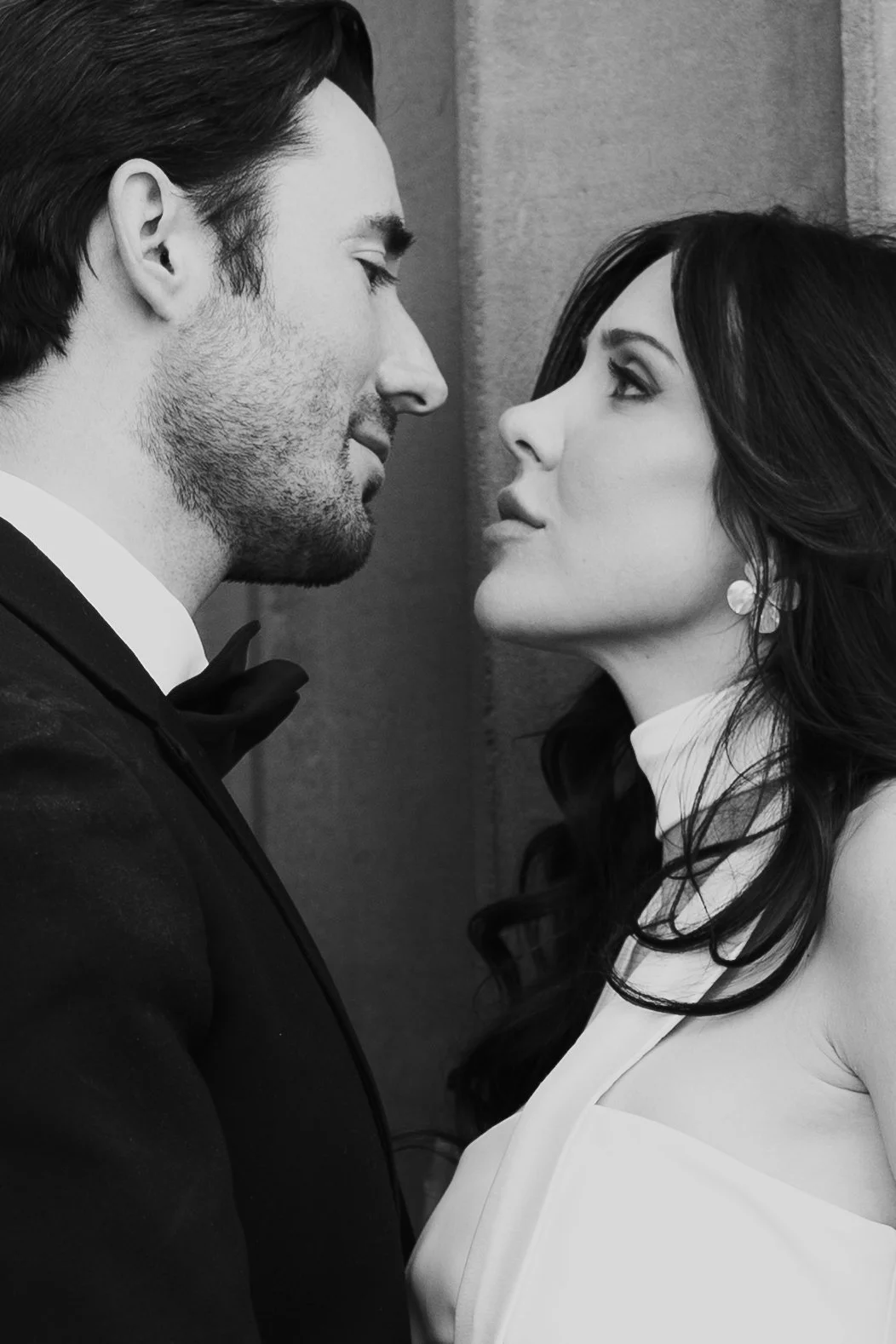 A bride and groom stand together among roman - style architecture in an elegant wedding gown and a classic black suit