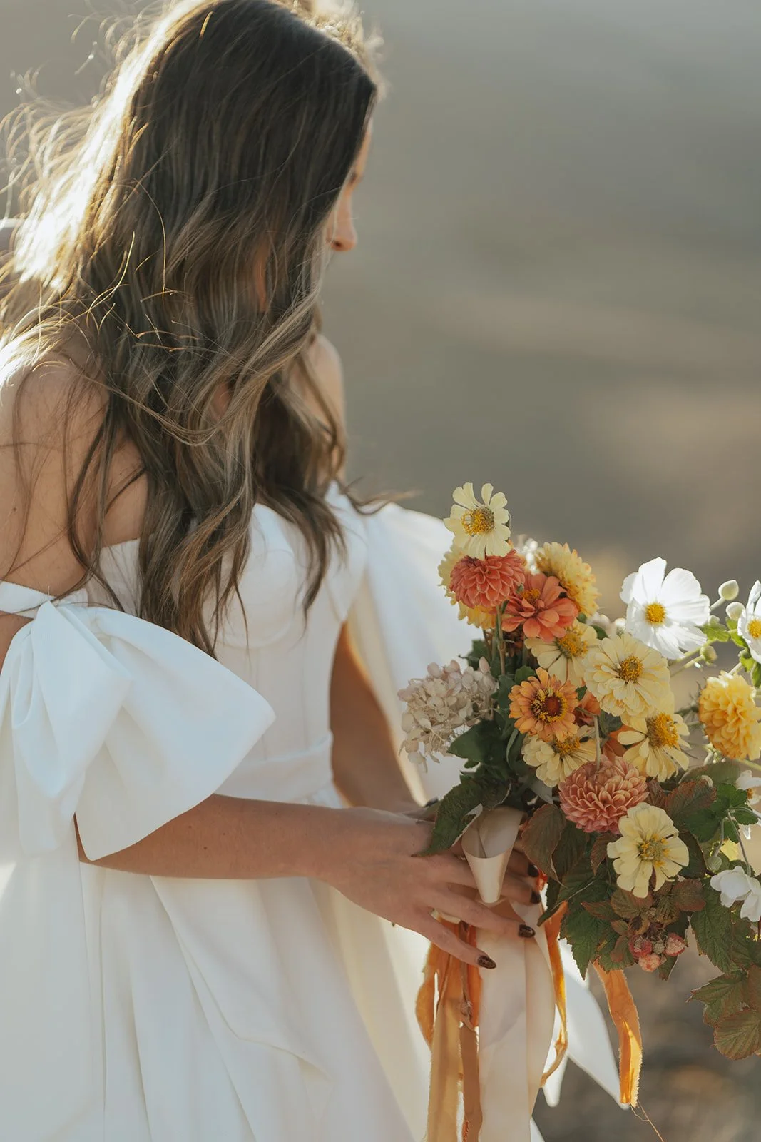 a bride wearing a wedding gown holds a bridal bouquet of white, yellow, and orange blooms as the sun provides a soft halo effect from behind her. Photography by Poise and Ivy Imagery