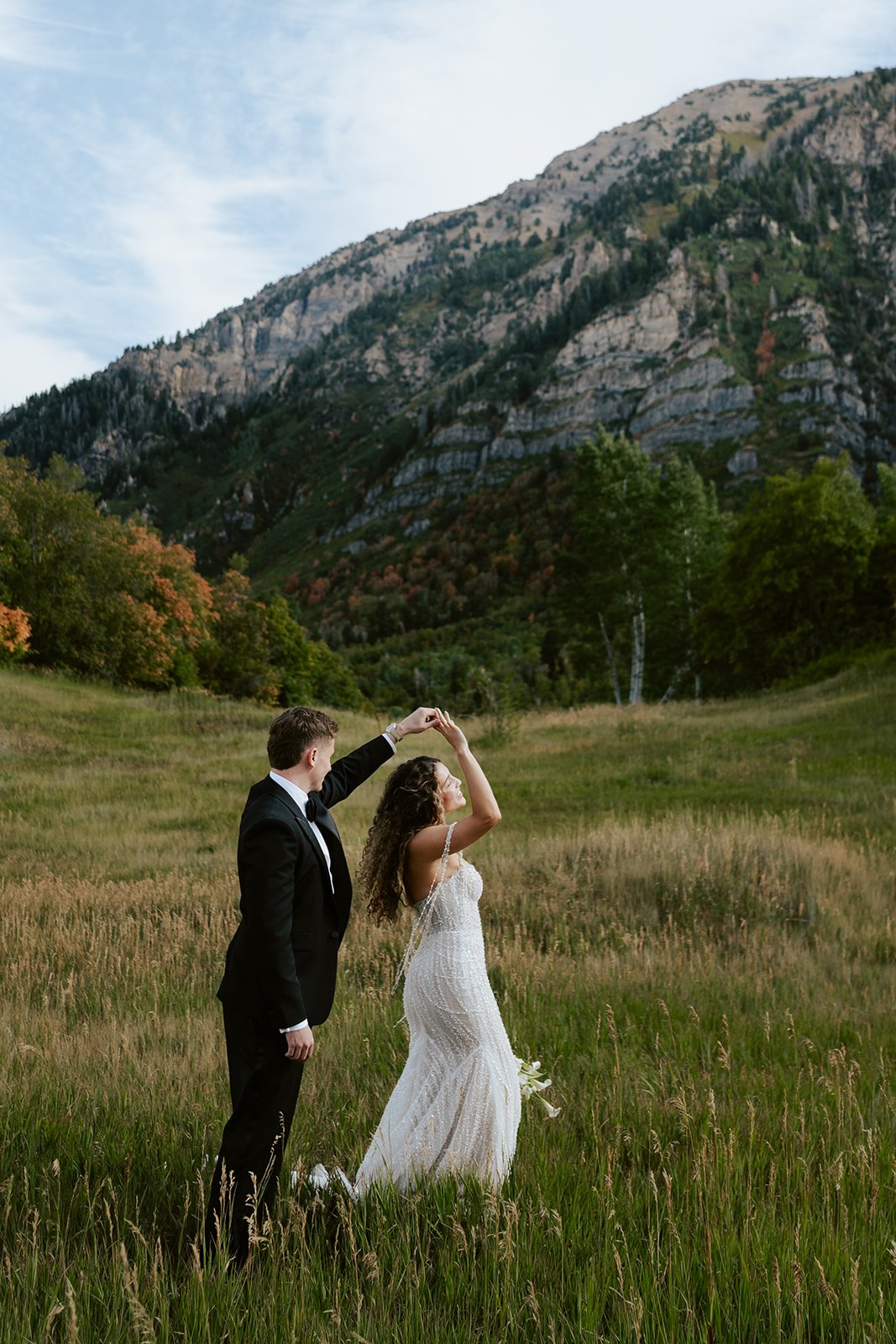 a bride and groom stand together in a softly sunlit meadow, wearing a beaded wedding gown and a black tuxedo. Photography by Poise and Ivy Imagery