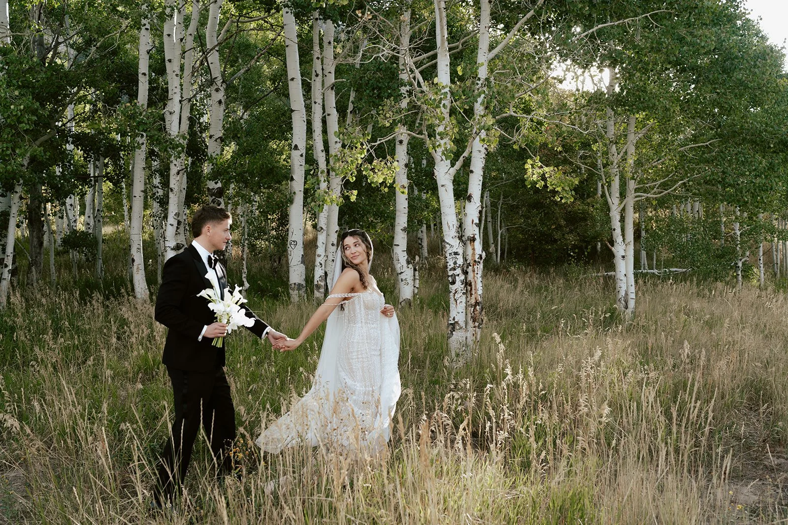 a bride and a groom walk together in a lush aspen grove. the bride wears a beaded wedding gown with a bridal veil and the groom wears a tuxedo. Photography by Poise and Ivy Imagery