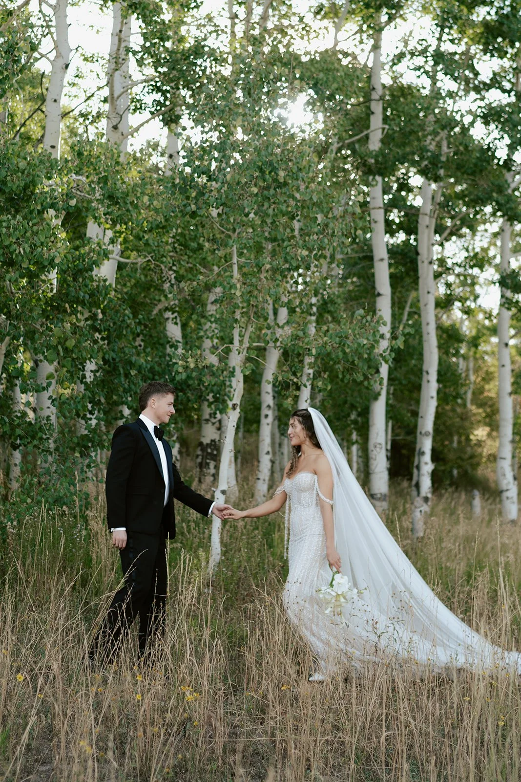 a bride and a groom stand together in a lush aspen grove. the bride wears a beaded wedding gown and the groom wears a tuxedo. Photography by Poise and Ivy Imagery