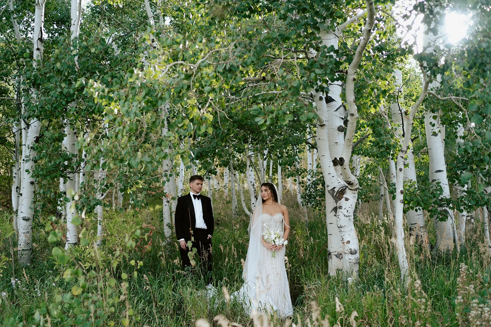 a bride and a groom stand together in a lush aspen grove. the bride wears a beaded wedding gown and the groom wears a tuxedo. Photography by Poise and Ivy Imagery