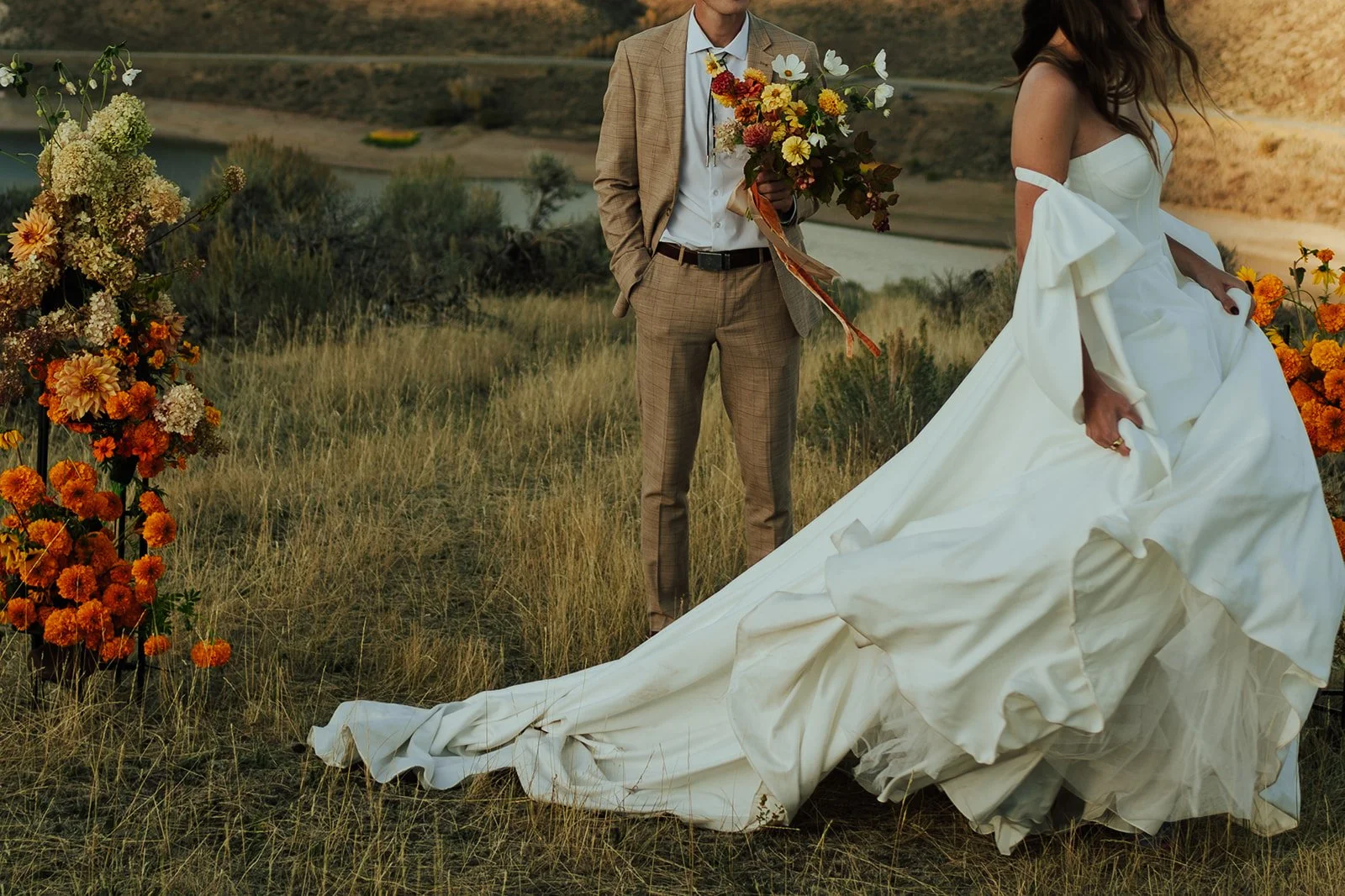 A bride and groom in a wedding gown and a tan suit stand in a meadow with mountains and a lake behind them - photography by Poise and Ivy Imagery