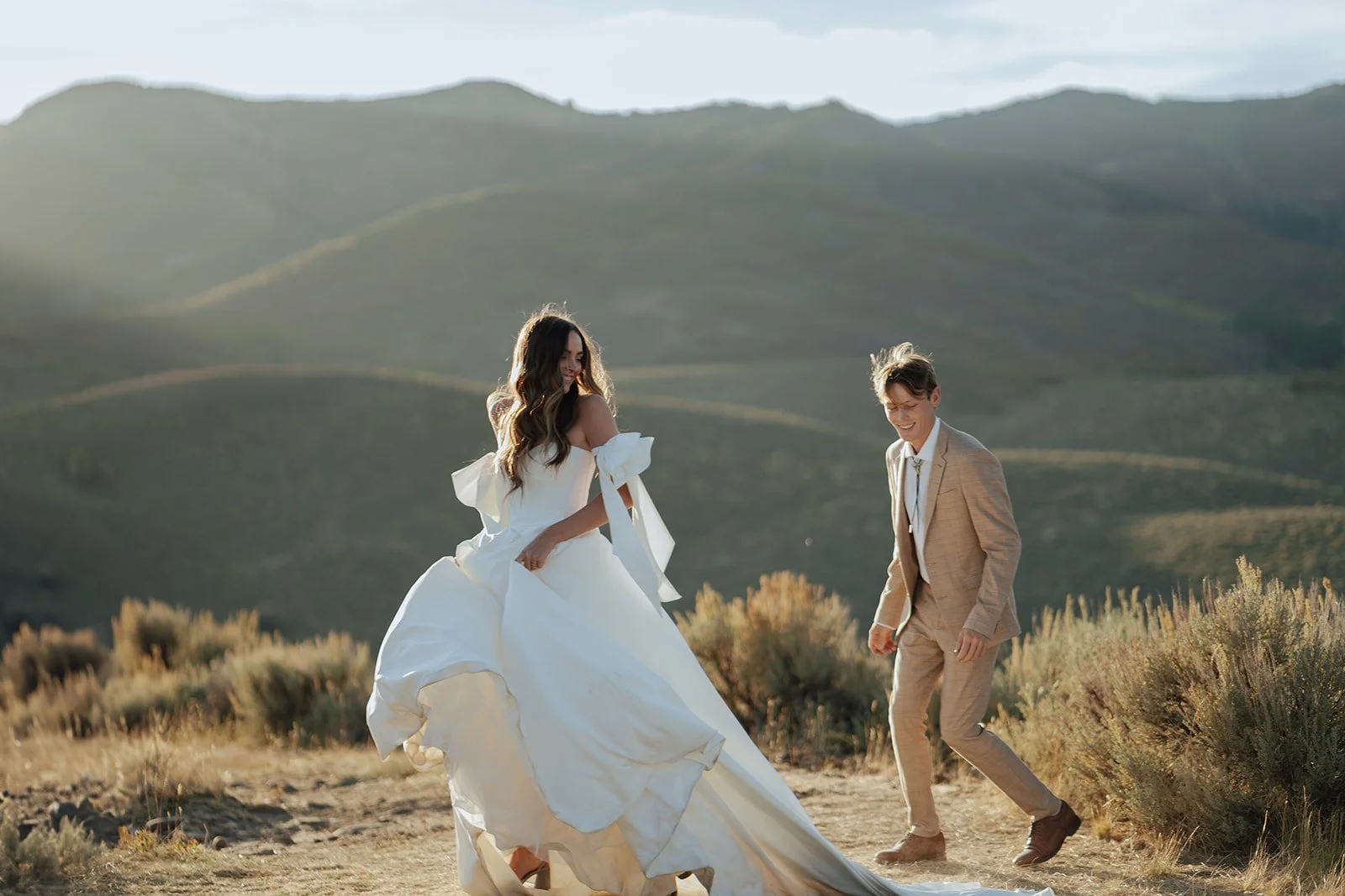A bride and groom in a wedding gown and a tan suit stand in a meadow with mountainous peaks behind them - photography by Poise and Ivy Imagery